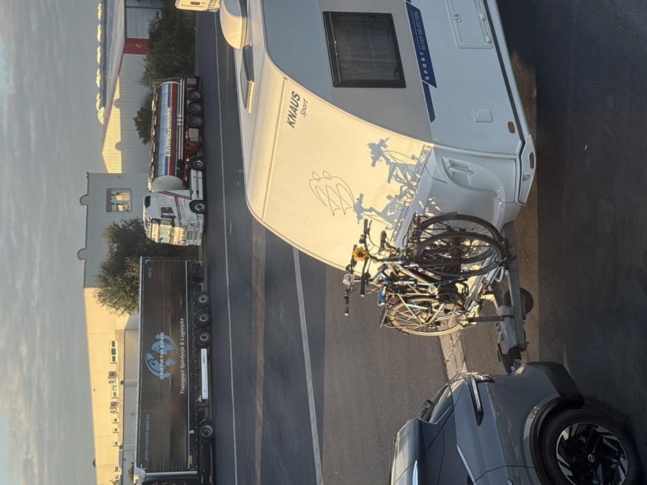 A grey crossover SUV is shown from the rear-left, hitched to a white Knaus Sport Silver Selection caravan. Two dark-colored bicycles are mounted on a rack attached to the tow hitch between the vehicles; a yellow sunflower is affixed to the handlebars of the front bicycle. The caravan features "KNAUS Sport" branding, a window on its left side, and a stylized graphic on the upper rear panel, illuminated by low-angle sunlight that casts elongated shadows of the bicycles onto the caravan's rear.
In the mid-ground, a dark semi-trailer truck with "CARPATIA SPED Transport Spedycja & Logistyka" branding is parked or stationary. To its right, a white semi-truck pulls a red, white, and blue tanker trailer. Behind these vehicles, industrial-style buildings with light-colored facades and various rooflines are visible under a partly cloudy sky. The entire scene takes place on a paved surface, likely a parking area or service road, in Himmelkron, Germany.