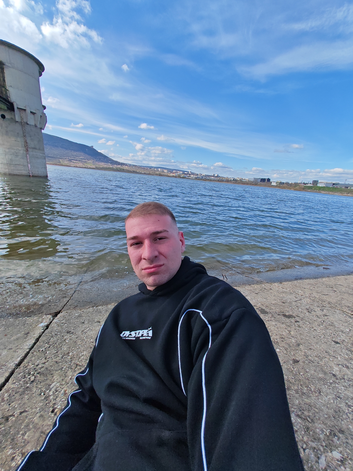 The photograph depicts a man sitting near a body of water. He is wearing a black hoodie with white accents. To the left, a concrete structure rises from the water. In the distance, a hill is visible, and further away, a city skyline can be discerned. The sky is blue with scattered clouds. The location is likely near the city of Shumen, Bulgaria, based on the terrain and architecture of the distant city. The water appears to be a lake or reservoir.