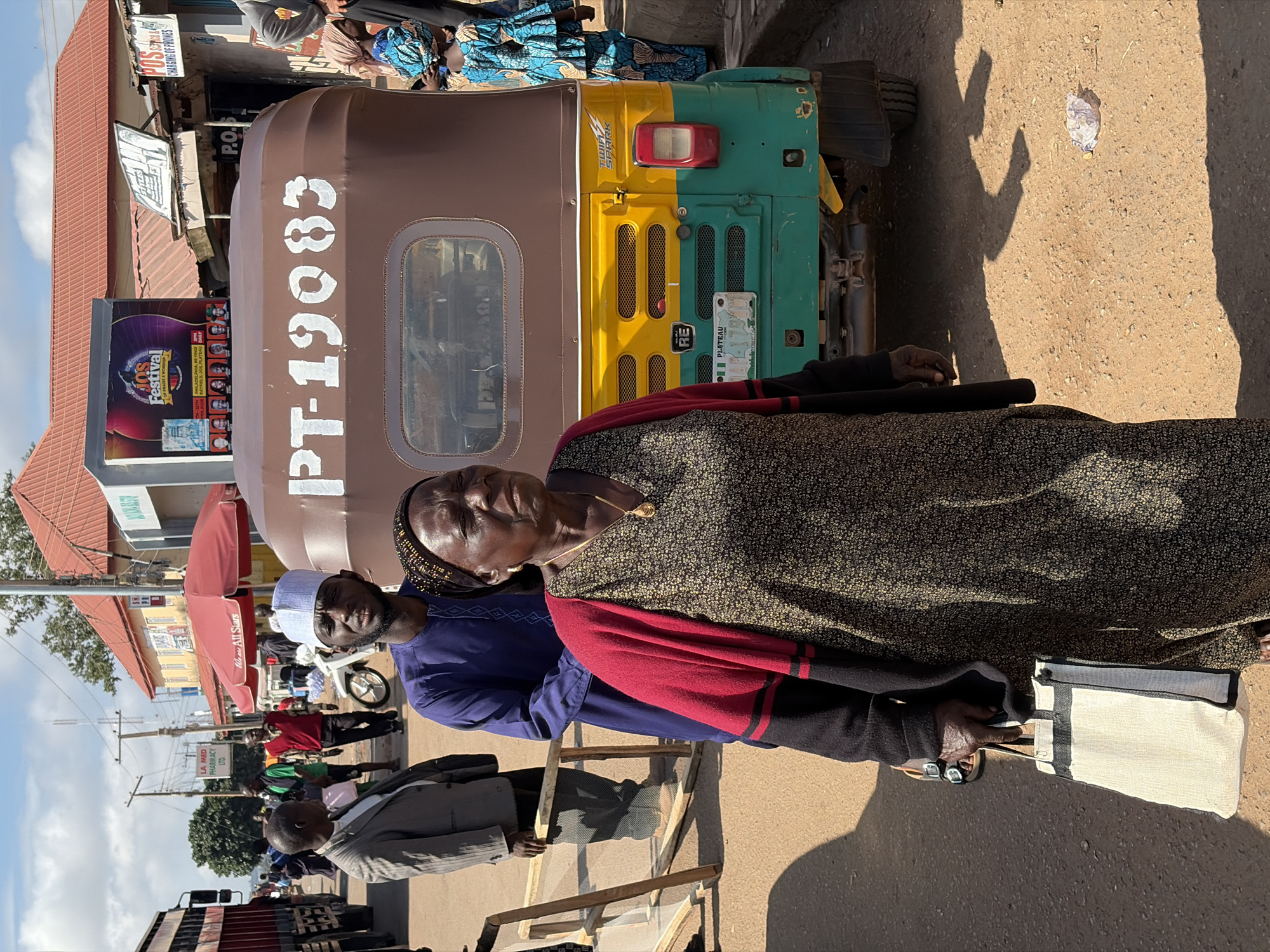 The image depicts a street scene in Gura Top, Nigeria, under a bright, sunny sky. In the foreground, an elderly woman with dark skin and wearing a patterned dress and a red shawl is looking to her right. She has a gold pendant on a necklace and is holding a light-colored tote bag in her left hand. To her left stands a man wearing a blue traditional outfit and a white cap, looking forward. Behind them, a three-wheeled motorized vehicle, commonly known as an auto-rickshaw, is parked. The vehicle is painted brown on the top half with white lettering "PT-19083" and a yellow and green lower half. Several other people are visible in the background, including men in suits and traditional clothing, and a woman in a vibrant blue and yellow patterned dress. The environment appears to be a bustling market or town square with buildings, utility poles, and signs, including one for "LA MED PHARMACY LTD." and another with "POS CHARGING OF PHONES." The ground is unpaved and dusty.