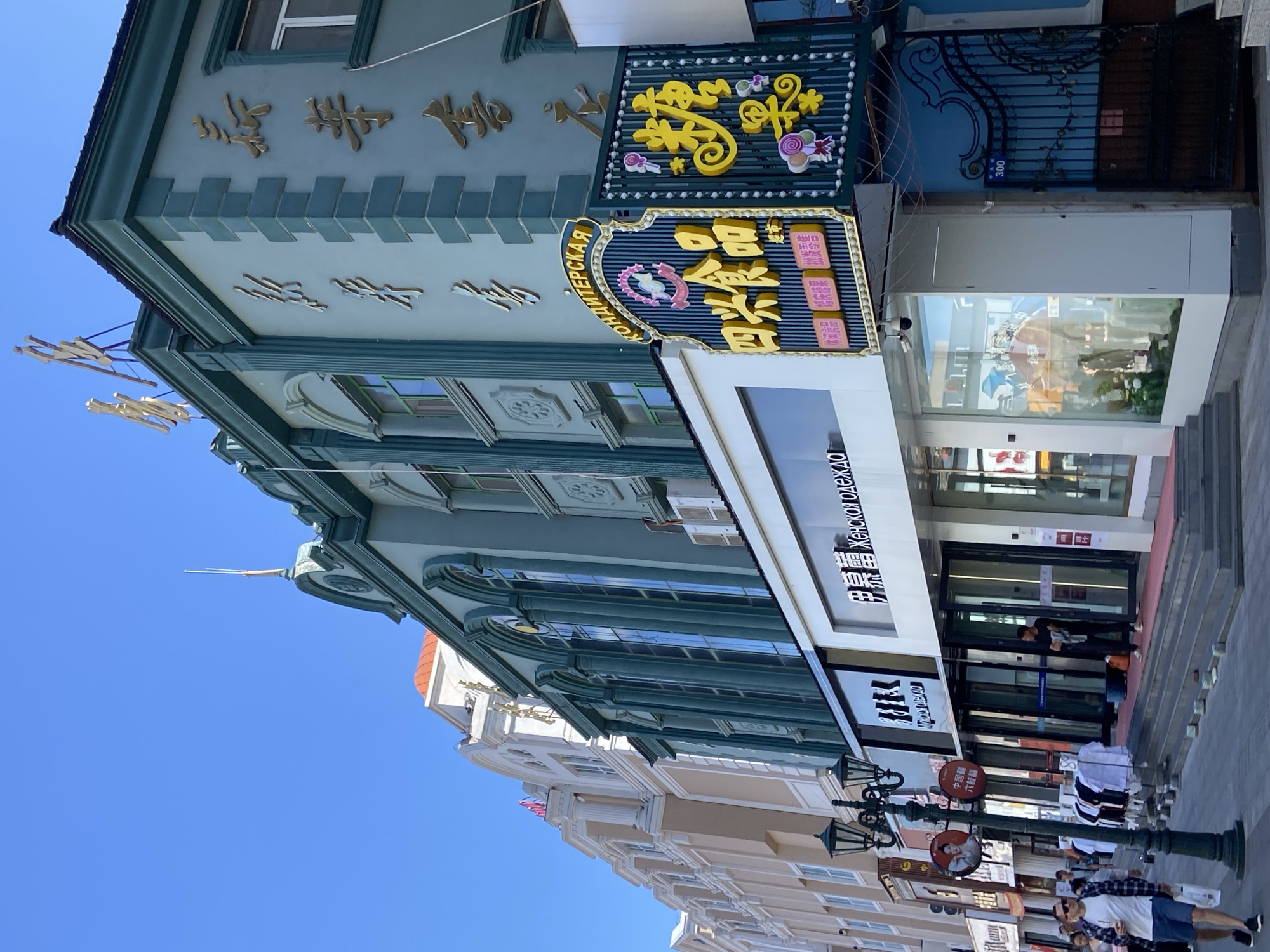 The image shows a street scene in Xingan Street, China. The buildings are styled with European architectural elements, featuring arched windows, columns, and ornate trim. The building on the right is painted a teal color with gold accents and large gold characters on its facade. A prominent sign with yellow Chinese characters and illustrations of candy hangs from this building. To the left, there are buildings in lighter colors, one appearing to be a pale yellow with white trim. A streetlamp with multiple lights stands on the sidewalk. Several shops are visible, with some displaying clothing on racks outside. People are walking on the sidewalk. The sky is clear and blue. The street appears to be a commercial area.