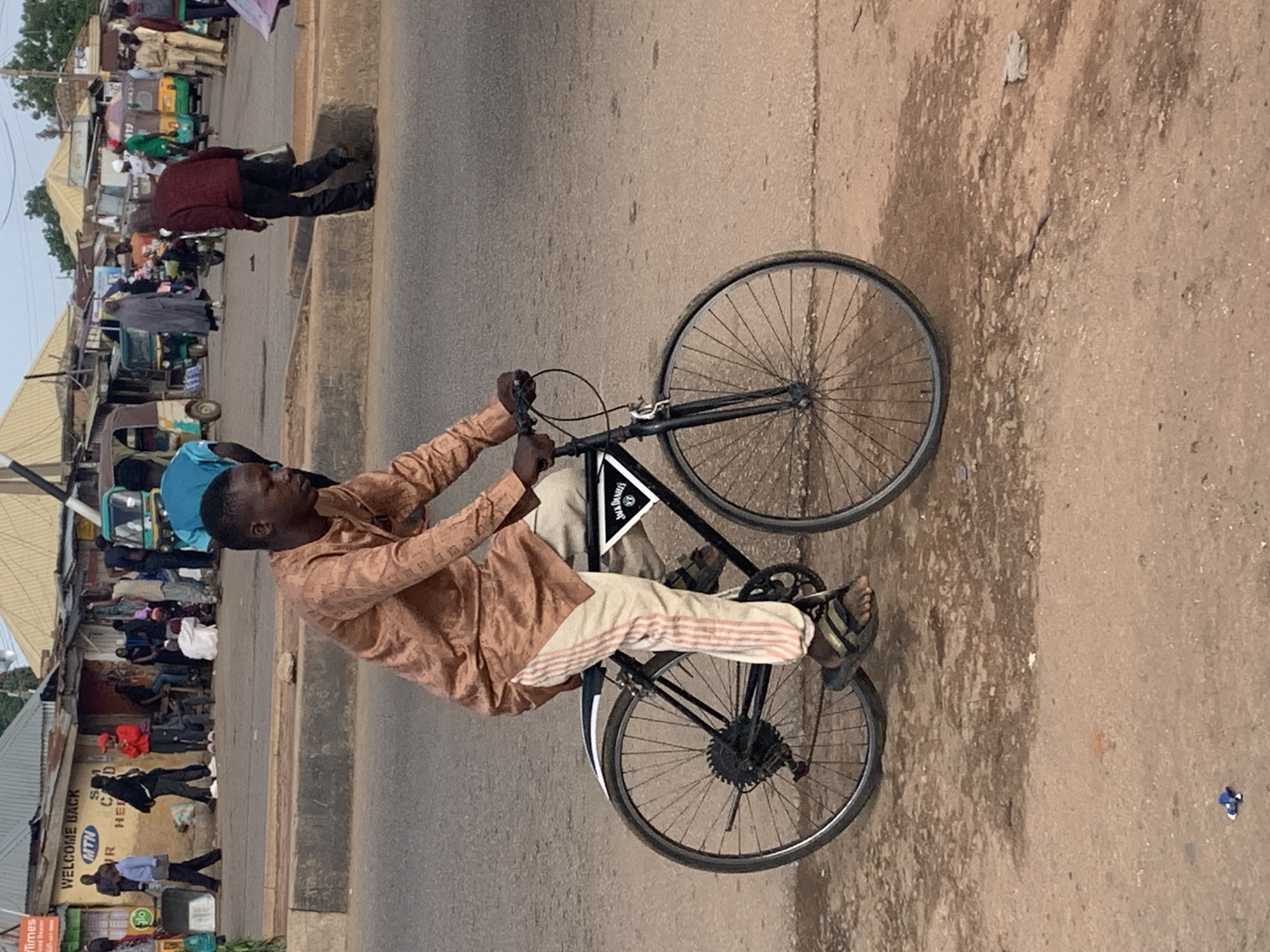 In Gura Top, Nigeria, a man in a patterned tan garment and striped pants rides a bicycle. He is wearing sandals. The bicycle has a black frame and fenders, and a small triangular decal with "Jack Daniel's" printed on it is affixed to the rear fender. The man is in motion, with his hands on the handlebars and his feet on the pedals. The background shows a street scene with several people, buildings, and vehicles, including what appear to be auto-rickshaws, indicative of a busy urban environment. The ground is dirt and appears somewhat worn.