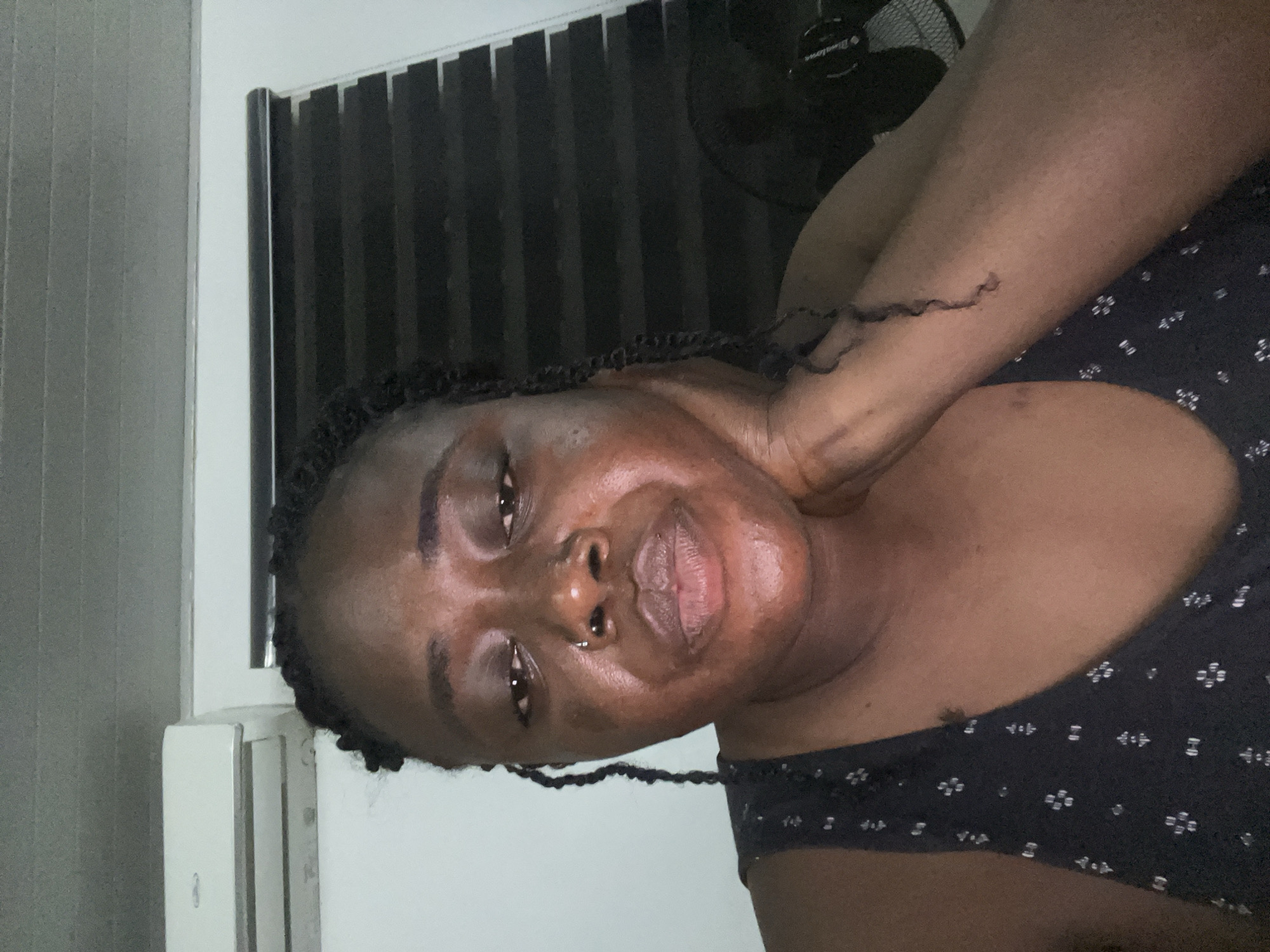 A close-up, overhead shot captures a dark-skinned woman with braided hair and a nose ring, resting her chin on her hand. The background features a white wall with an air conditioning unit mounted near the ceiling on the left, and black horizontal blinds on the right. The lighting is dim, casting subtle shadows. The scene appears to be indoors, possibly a bedroom or living area, in Abuja, Nigeria.
