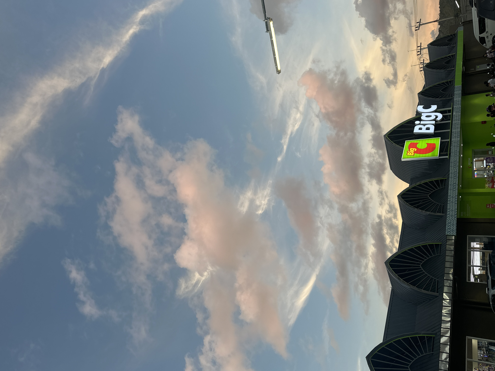 This media file captures a low-angle view of the sky and the exterior of a "Big C" supermarket in Ko Pha Ngan, Thailand. The sky is a prominent feature, filled with a mix of light blue and scattered clouds, some of which are tinged with pink and orange, suggesting either sunrise or sunset. Wispy clouds streak across the upper portion of the image, while more voluminous, textured clouds are present lower down. A long, fluorescent light fixture is visible against the sky in the upper left quadrant, attached to a pole.
The supermarket building occupies the right side of the frame. It has a modern architectural design with a dark, curved roof structure that is segmented by fan-like awnings over windows. The prominent "Big C" logo is displayed in large, illuminated white letters against a dark background, with a smaller, green illuminated sign featuring the "Big C" logo to its left. The building's facade is predominantly green and dark gray. Visible through windows are displays and what appear to be aisles within the store. A white van is parked to the far right, and there are hints of people and motorcycles near the entrance.
The overall impression is a snapshot of a commercial establishment in a tropical setting, with the natural beauty of the sky providing a dramatic backdrop. The time of day appears to be either early morning or late afternoon/early evening, given the colors in the sky. The weather seems fair with scattered clouds. The visible text includes the "Big C" logo.