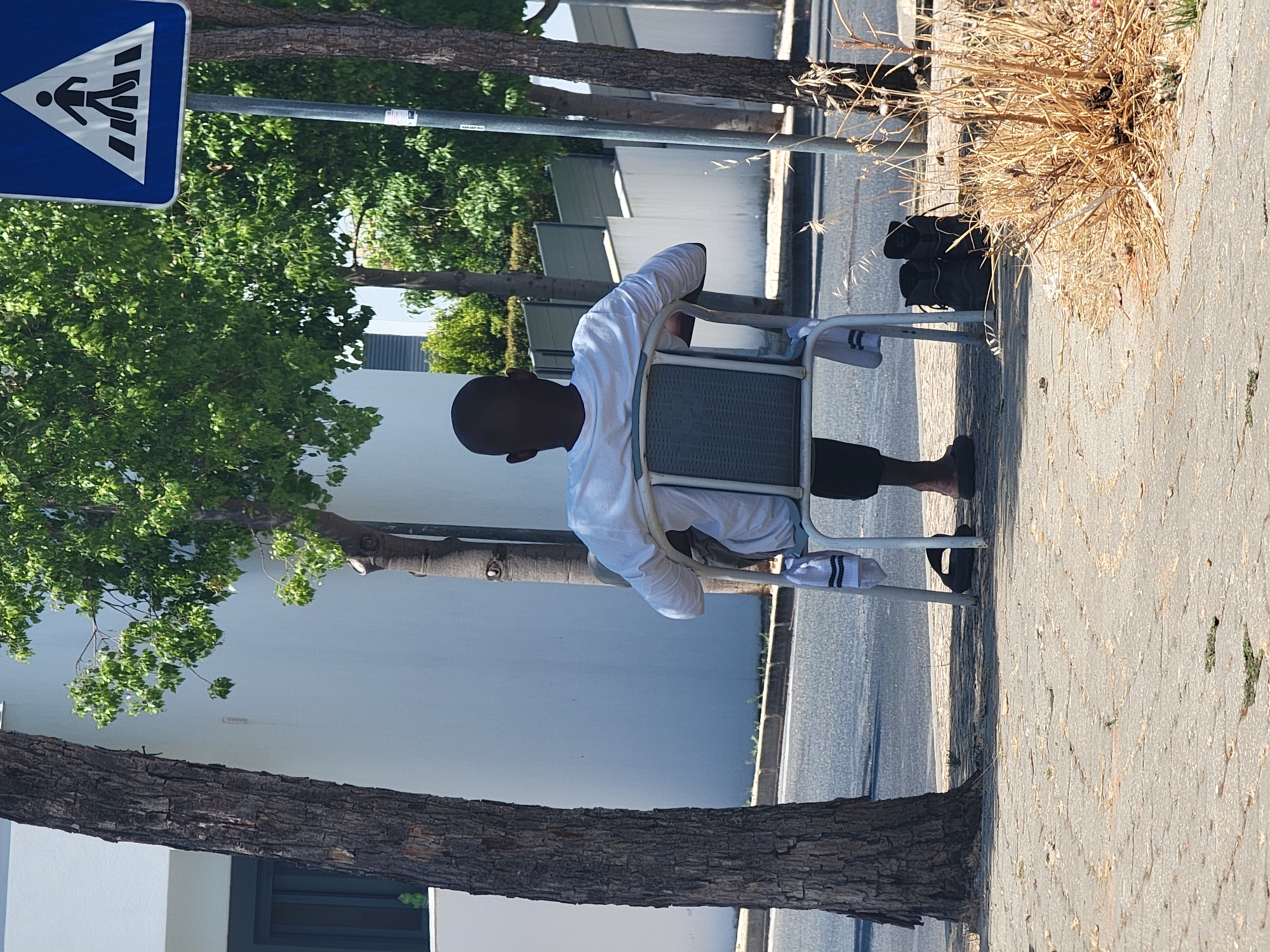 A Black male is seated in a chair facing away from the camera. He is wearing a white long-sleeved shirt and dark pants. A pair of black sandals and black boots are on the ground near the chair, along with a white sock. The scene takes place outdoors on a sidewalk next to a street, with trees and a white building in the background. A pedestrian crossing sign is visible to the right of the image. The setting appears to be a city, possibly Quarteira, Portugal, given the style of the building and the presence of a pedestrian crossing sign.