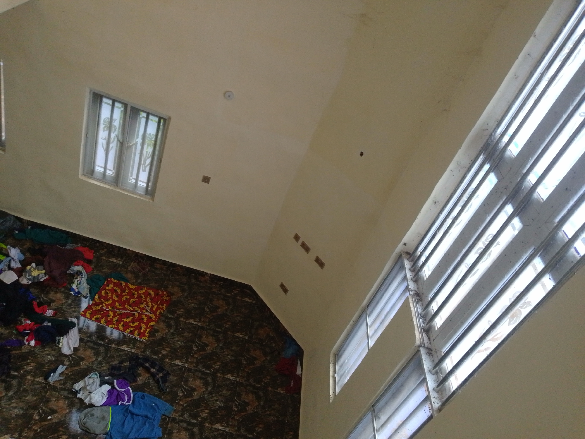 The image depicts an interior scene, likely within a residential building in Obosi, Nigeria. The perspective is from a high angle, looking down onto a tiled floor and up towards a sloped ceiling.
On the floor, scattered clothing items are visible. These include a red and yellow patterned cloth, various t-shirts, shorts, and other apparel in different colors such as blue, purple, grey, and black. Some items appear to be folded, while others are in disarray. The floor itself is covered in dark, glossy tiles with a marbled pattern.
The walls are a light, neutral color. A window with vertical bars and reflective glass is situated on one wall, allowing light to enter the room. Another window, also with horizontal blinds, is visible on a higher plane, suggesting multiple levels or an open-plan design. The ceiling is a pale, cream color and shows some signs of minor imperfections or water stains in places. Several square ventilation grates or electrical boxes are embedded in the ceiling. In the upper part of the image, a circular object, possibly a smoke detector or light fixture, is mounted on the ceiling. The overall impression is of a somewhat untidy but functional living space.