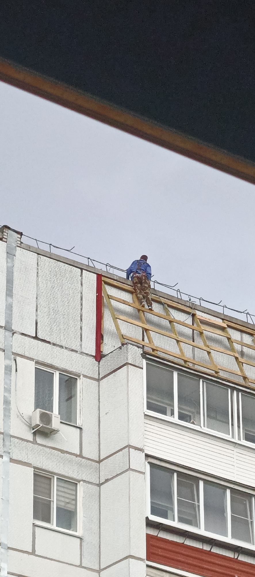 The image displays the upper section of a multi-story building, likely residential, under an overcast sky. A single person, dressed in a blue jacket and camouflage-patterned pants, is positioned on the building's rooftop or top edge. They appear to be engaged in work, possibly climbing or working on a wooden frame structure that is being constructed on the top of the building. This wooden frame extends horizontally across the facade, suggesting ongoing construction or renovation, possibly for a new roof or an extended section.
The building itself is constructed from grey concrete panels, a common architectural style. Protruding metal rods are visible along the top edge of the concrete structure. A distinctive red vertical element is present on the left side of the wooden frame. Below the work area, an air conditioning unit is mounted on the building's exterior wall, beneath a window with a white frame. Other windows are also visible, some showing white horizontal blinds. The very top of the image is partially obscured by a dark, out-of-focus object, possibly an overhang or a railing from another vantage point. The scene depicts a construction or maintenance activity during daytime under cloudy weather conditions. There is no discernible text in the image.
