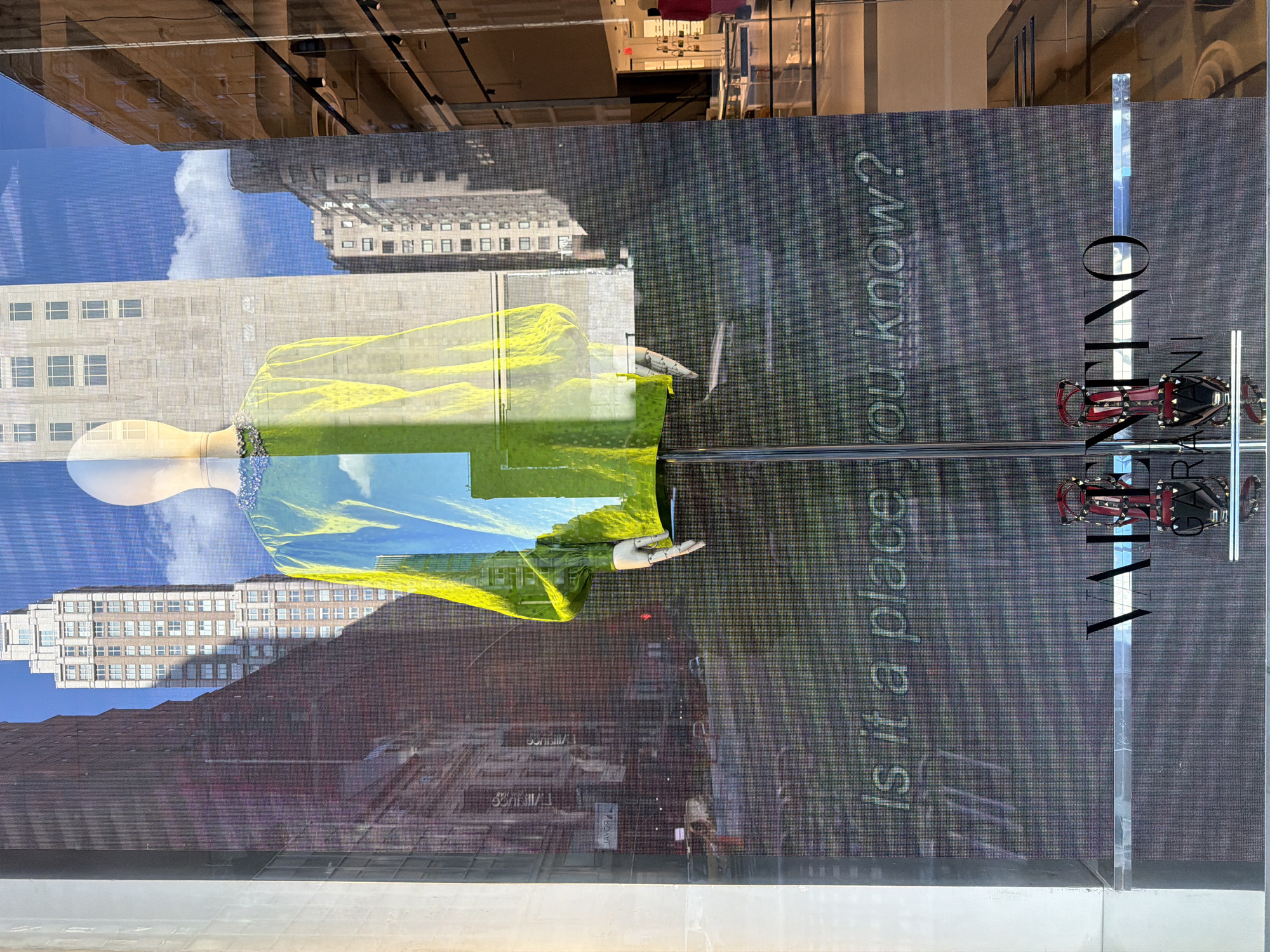 This is a photograph of a store window display in New York City, United States of America, likely on Fifth Avenue or a similar upscale shopping district. The display features a mannequin dressed in a bright, translucent yellow and blue garment adorned with embellishments around the neckline. The mannequin is positioned in front of a digital screen that projects an image of the New York City skyline, complete with tall buildings and a bright blue sky with clouds. Below the mannequin, text that reads "Is it a place you know?" is displayed on the screen. At the bottom of the display, a pair of red, studded high-heeled shoes are showcased next to the "VALENTINO" logo. The overall scene conveys a sense of luxury and urban sophistication, typical of high-end fashion retail in New York City.