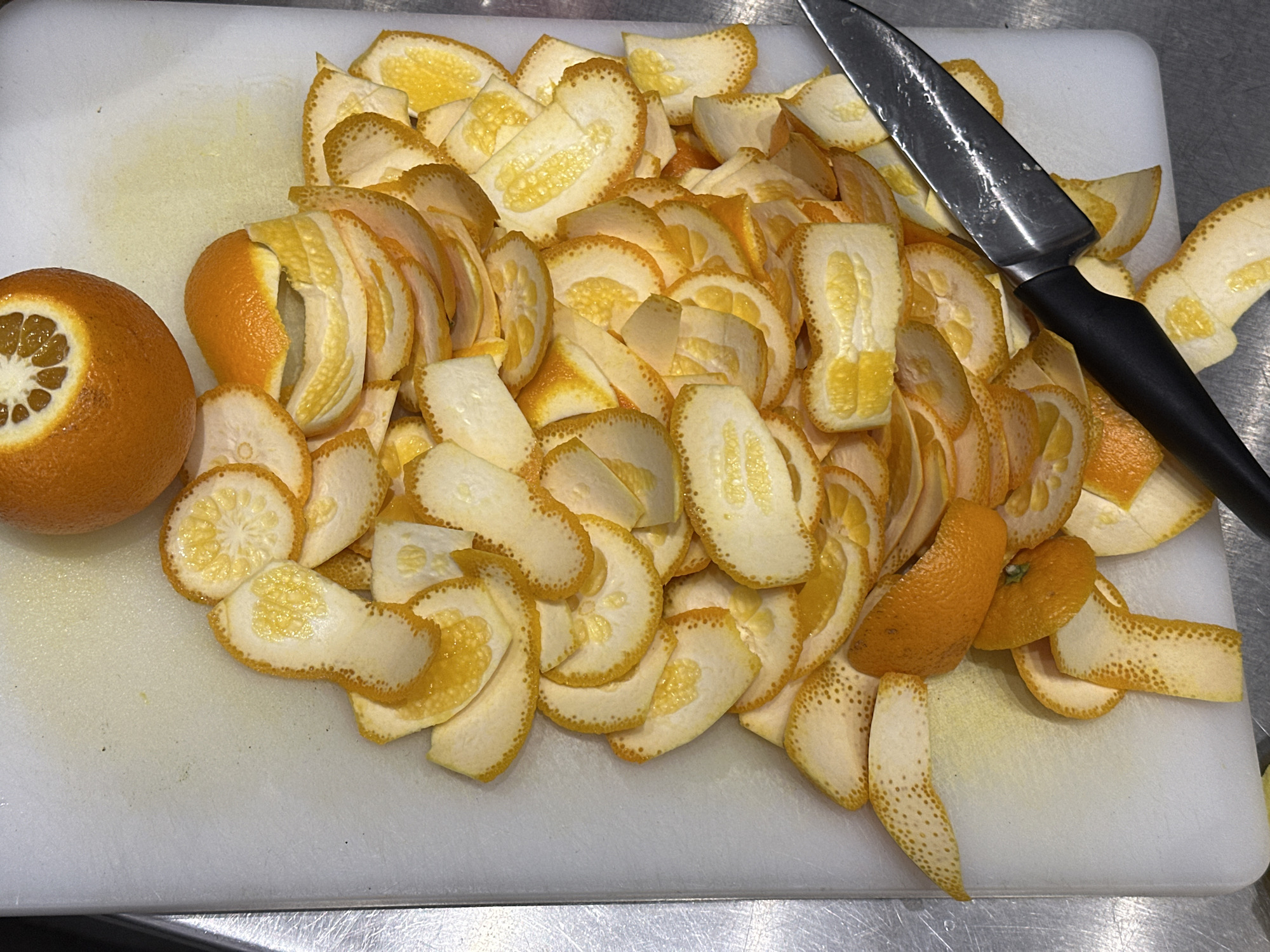 A white cutting board on a stainless steel surface is piled with numerous segments of orange peels, each partially retaining the yellow, spongy pith, and occasionally with small sections of the orange skin still attached. An orange fruit, halved, sits on the side, showing its internal segments. A knife with a black handle and a silver blade rests on the board amongst the peels. The setting appears to be a kitchen. No information about Novi Sad, Serbia, is apparent from the image.