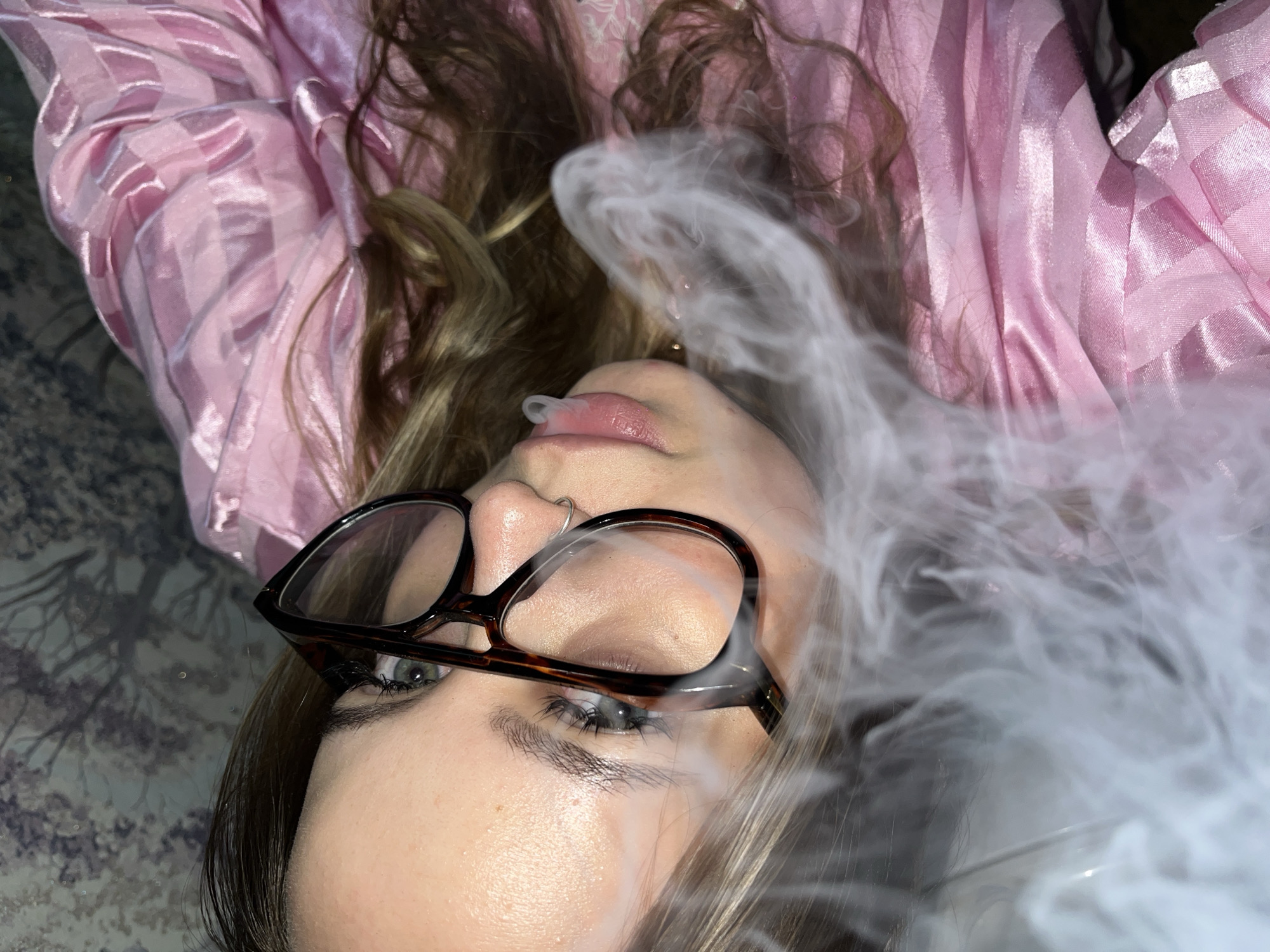 A person lies on their back, wearing glasses and a pink patterned top. Smoke emanates from their mouth. The location is unspecified.