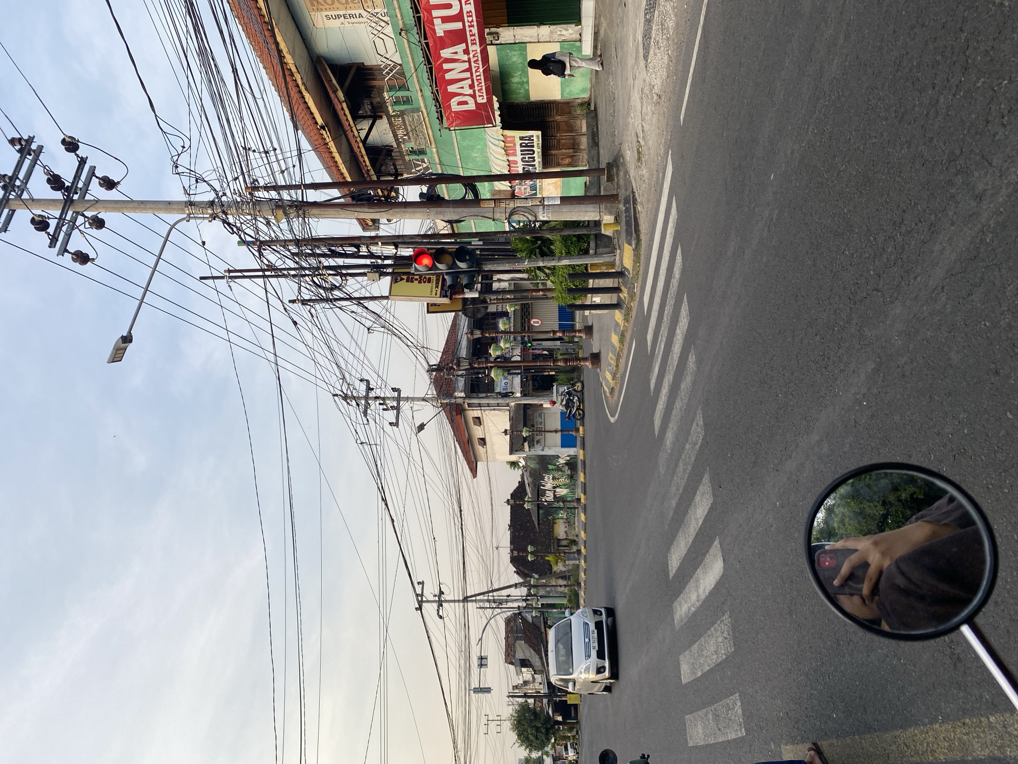 The image depicts a street scene in Madiun, Indonesia, taken from the perspective of a motorcyclist stopped at a crosswalk. A white car with license plate AE 1419 BM is visible ahead. Above, utility poles covered in a tangle of wires stretch along the street. Buildings line the street, including one with signage for "DANA TU," "JAMINAN BPKB M," "SUPERIA," and "TTO KILAT PIGURA." A traffic light is showing red. The side mirror of the motorcycle reflects the motorcyclist's hand holding a cell phone.