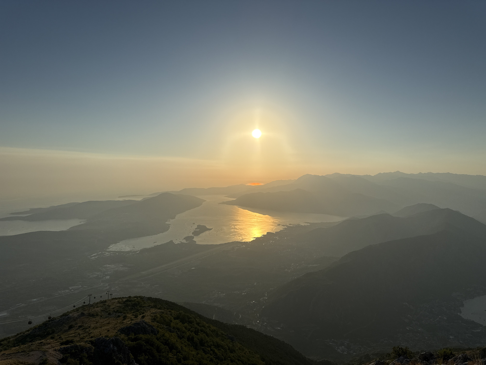 The image is a panoramic view of a mountain range overlooking a bay at sunset, likely from the peak of Mount Vrmac near Kuk, Montenegro. The sun, a bright, intense orb, hangs in the sky, casting a golden-orange glow that reflects on the water. Layers of hazy, silhouetted mountains recede into the distance, creating a sense of depth. In the foreground, a rocky, grass-covered slope descends towards a body of water. A cable car system with several gondolas is visible on the left side of the frame, traversing the mountainside. The water itself appears to be part of a large bay, with a town or village nestled along its shores, illuminated by the fading sunlight. The overall atmosphere is serene and majestic, capturing the natural beauty of the coastal landscape at dusk.
