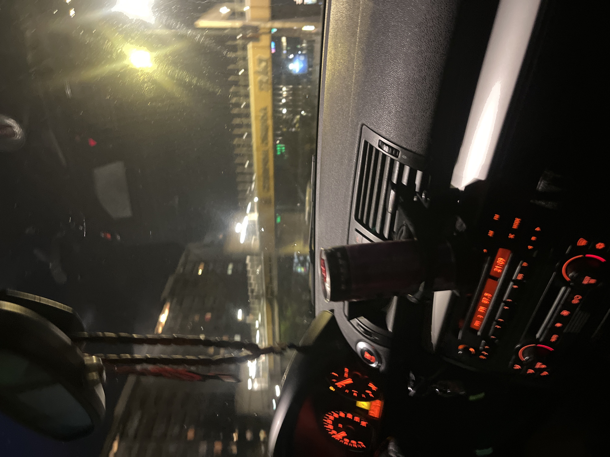 The media file captures an interior view of a car at night, looking out through the windshield onto a city scene. The perspective is tilted, suggesting the image might have been taken from a passenger's or driver's side with the phone held at an angle.
Inside the car, a portion of the dashboard and center console are visible on the right. The car's infotainment system or radio is illuminated with red/orange backlighting. The display clearly shows "21:09," indicating the time, and "FM 97.9," a radio frequency. Buttons labeled "AM," "FM," "MODE," and "AUTO" are also discernible. An air vent is prominent on the dashboard, and a red beverage can, possibly an energy drink, is clipped into one of the vents. Part of the steering wheel is seen in the lower left, also with red illuminated controls. Hanging from what appears to be the rearview mirror are decorative beads or a rosary, extending towards the lower left of the frame.
Outside the car, through the windshield, a vibrant urban environment in Sofia, Bulgaria, is illuminated by numerous artificial lights. Tall buildings with many windows, reflecting the city lights, line the streets. A prominent tall, yellow vertical structure with unreadable text is visible amidst the buildings. Streetlights and other vehicle lights create streaks and glows across the dark nighttime sky. The windshield shows some reflections, possibly from interior lights or condensation.
The overall impression is of being inside a vehicle, possibly stationary or slowly moving, during the evening in a busy city.