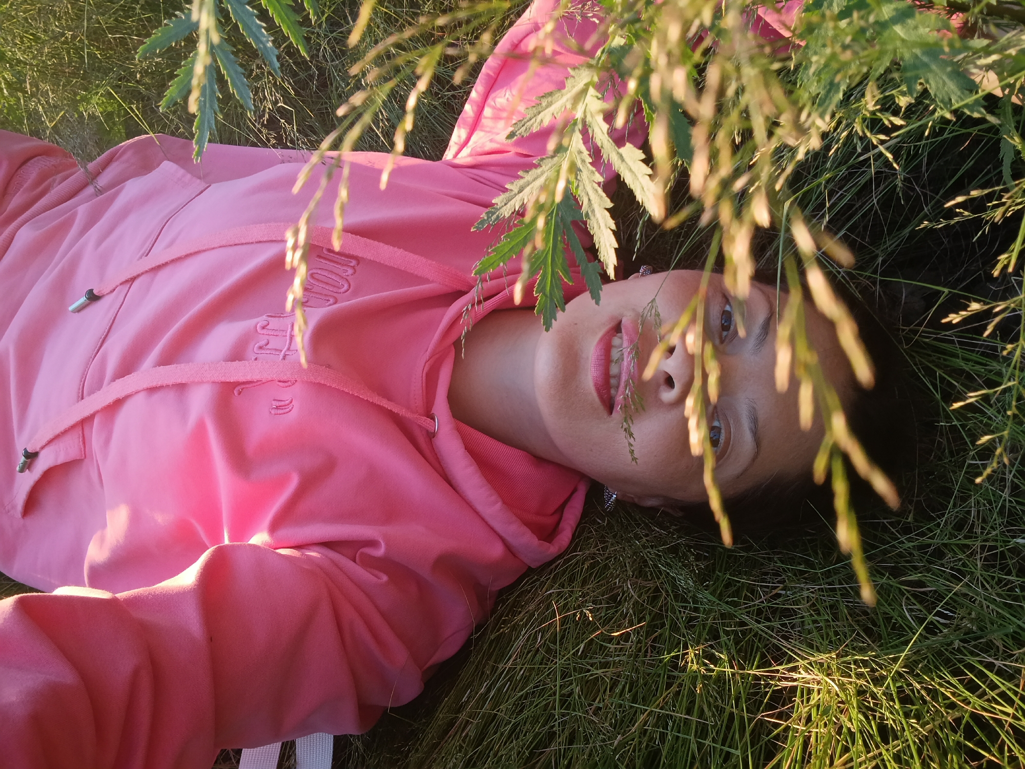 A woman in a pink hooded sweatshirt lies in the grass, her head propped up by her arm. Blades of grass and leaves partially obscure her face and the front of her sweatshirt. The woman appears to be looking at the camera, with her mouth slightly open, revealing her teeth. The lighting suggests it is either early morning or late afternoon. The image does not contain any discernible landmarks or features that would indicate a specific location within the city of Saratov, Russia.