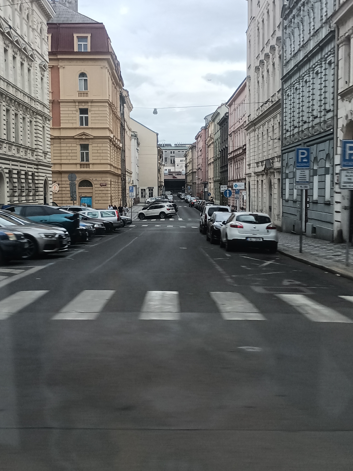 The image shows a street in Prague, Czechia, with buildings lining both sides of the road. The street is paved and has pedestrian crosswalks. Several cars are parked on the side of the street and a few are in motion. There are also some pedestrians visible. The buildings are of various architectural styles and appear to be old, characteristic of a European city. The sky is overcast.