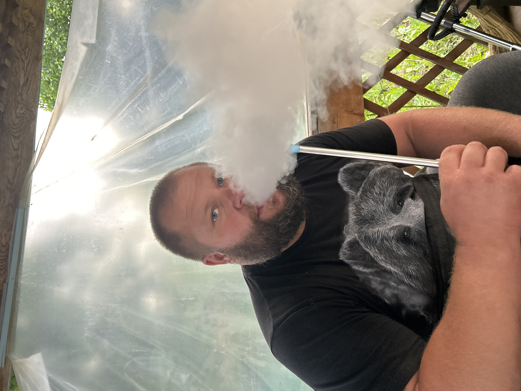 A man with a beard is exhaling a cloud of smoke. He has short brown hair and blue eyes, and is wearing a black T-shirt with a gray wolf image. He is holding a metal tube. The background includes translucent plastic sheeting and a wooden lattice with green foliage visible through it. The location is Фоки, Russia.