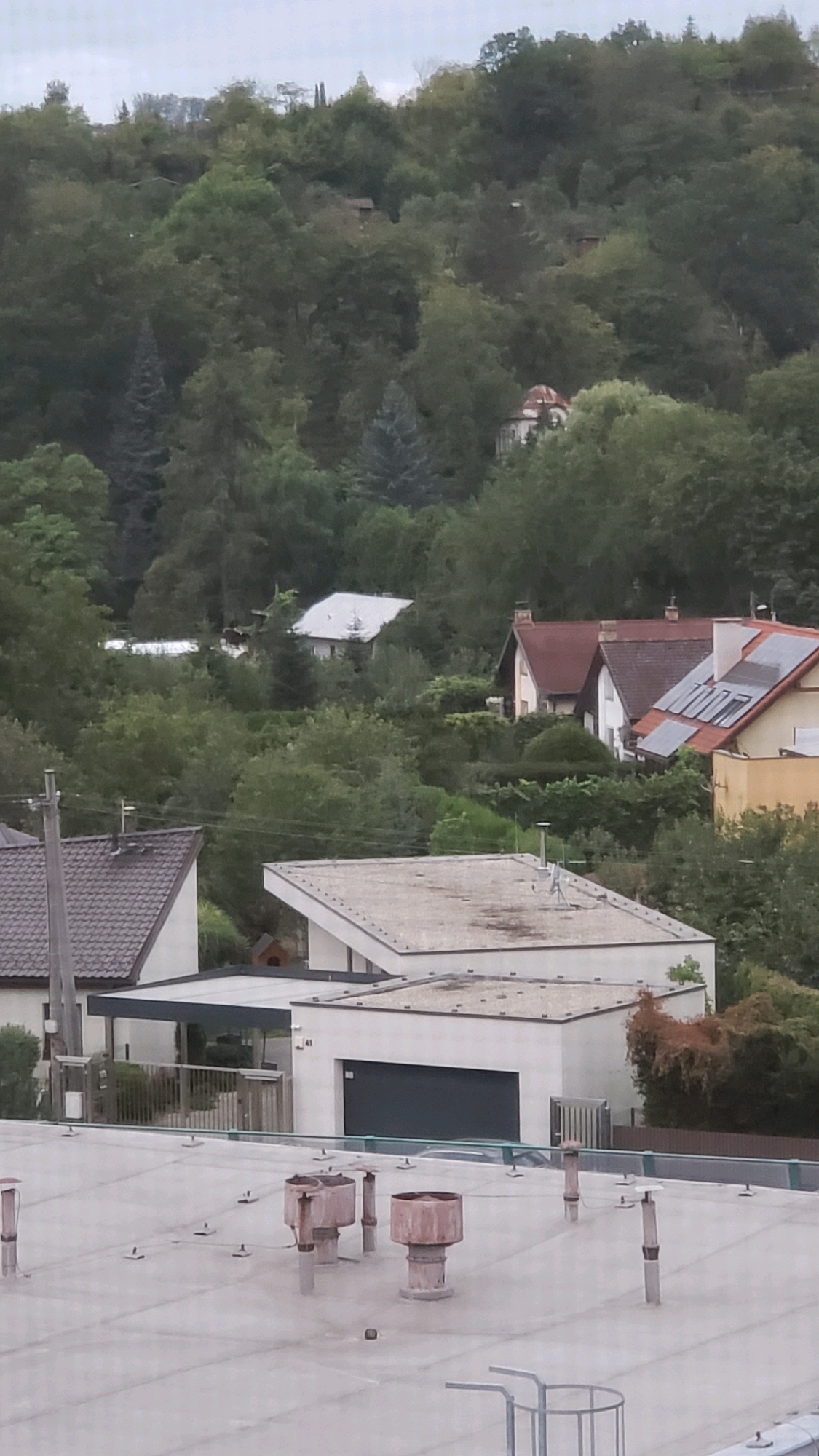 The image captures a view of residential buildings nestled within a wooded hillside, likely in Prešov, Slovakia, given the provided context. In the foreground, a flat, light-colored roof occupies the bottom of the frame, featuring several weathered vent pipes and some mounting hardware. Behind this roof, several modern structures are visible. A white building with a sloped roof and a dark garage door is situated centrally. To its left, another building with a dark-tiled roof and a grey pole is partially visible. Further up the hill, several houses with pitched roofs, some with visible solar panels, are interspersed among the dense green foliage. The background is dominated by a thickly wooded slope under an overcast sky. No people are present in the image.