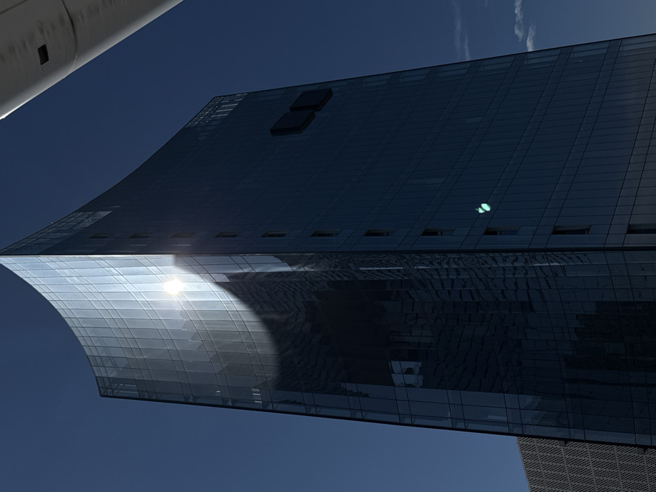 The low-angle image showcases a modern glass building against a clear blue sky. The building's architecture features a curved facade reflecting the sunlight, creating a bright glare. The rest of the building is dark blue with rectangular windows. A white object is at the upper left edge, possibly a part of another structure or vehicle. The architecture style is sleek and contemporary.
