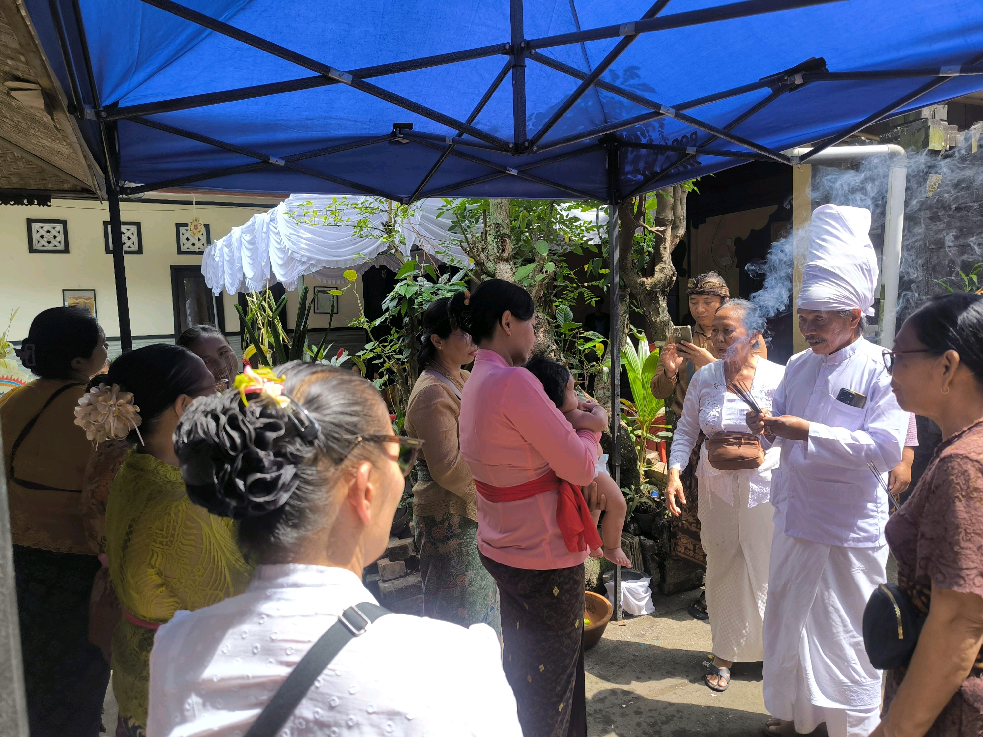 In Tabanan, Indonesia, a group of people is gathered under a blue canopy. The scene appears to be a religious or cultural ceremony. In the foreground, several women are seen from behind, one wearing a white shirt and another a green patterned garment. A woman in the center is holding a baby. To the right, a man dressed in white, including a distinctive head covering, holds incense sticks. He is standing next to an older woman who also appears to be holding incense. Smoke is rising from the incense, adding to the atmospheric feel of the scene. The background shows traditional Indonesian architecture with white decorative elements and lush greenery. The overall impression is one of a community event, possibly a prayer or a ritual, taking place outdoors in what seems to be a courtyard or a communal gathering space.
