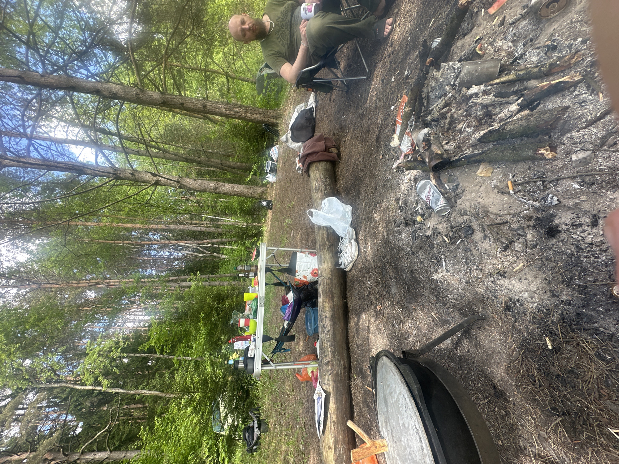 The image depicts a campsite in a wooded area. A bald man wearing a green shirt and dark pants sits in a folding chair, holding a container. A table laden with supplies is visible. A blackened fire pit containing charred wood, cigarette butts, and discarded cans is prominent in the foreground. A large cooking pot with a lid sits next to the fire pit. There's a fallen tree on the ground, and litter is scattered around. The location is likely in Юзуфоўскі сельскі Савет, Belarus, given the prompt.