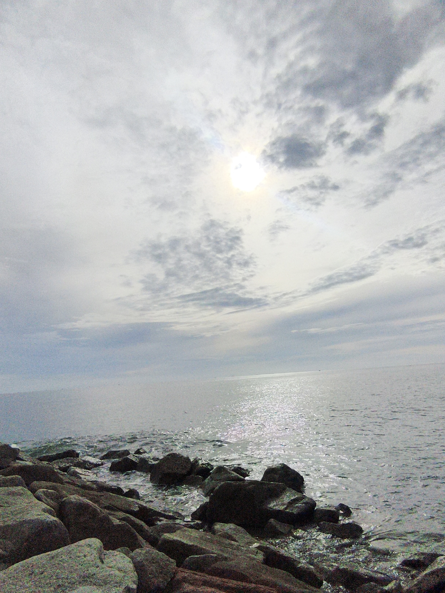 The image depicts a coastal scene with a rocky shoreline in the foreground, leading to a calm body of water. The water is reflecting the light of the sun, which is partially obscured by clouds in the sky. The sky is a mix of white and gray clouds, with the sun creating a bright, hazy glow. The water appears still and tranquil, with small ripples on its surface. The rocks are varied in size and shape, some appearing smooth and rounded, others more angular and jagged. Some of the rocks are wet from the water lapping at the shore. The overall impression is one of a quiet, natural landscape. This scene could potentially be located along the coast of Lhokseumawe, Indonesia, which has numerous beaches and rocky shorelines.