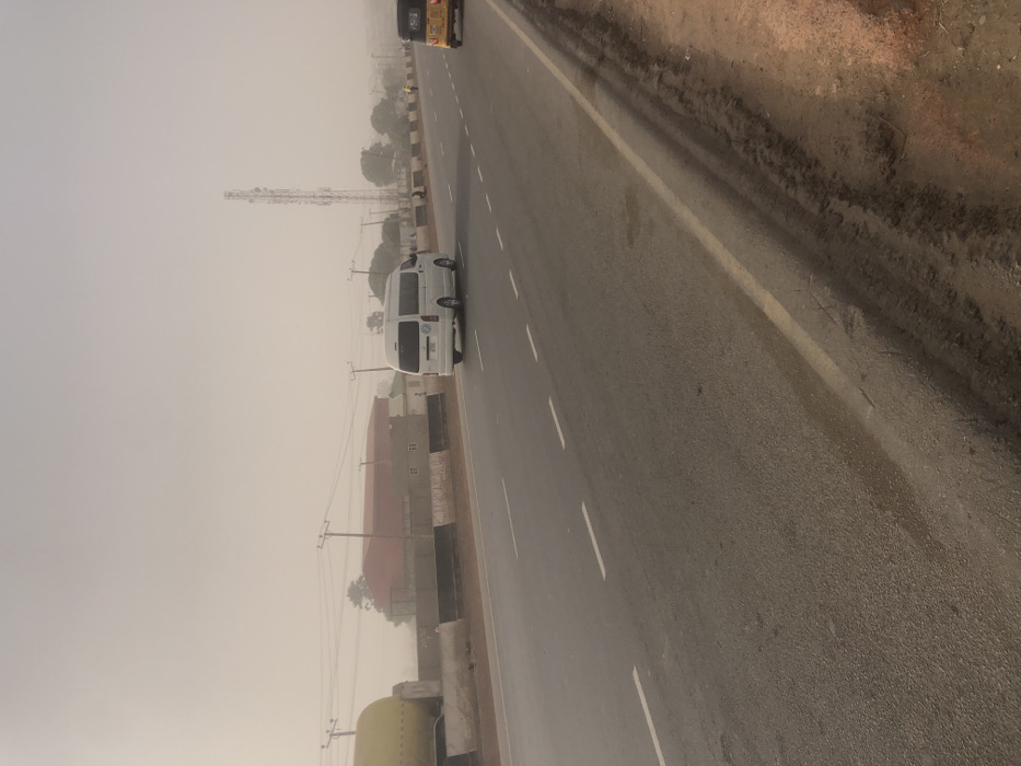 A hazy road view features a white van traveling on the left lane. A yellow vehicle is partially visible in the top right corner. Power lines run parallel to the road. A cell tower stands in the distance on the left. Adjacent to the road are buildings with red roofs, partially obscured by the haze. The sky is overcast, creating a muted color palette. Road markings are visible, indicating lane divisions.