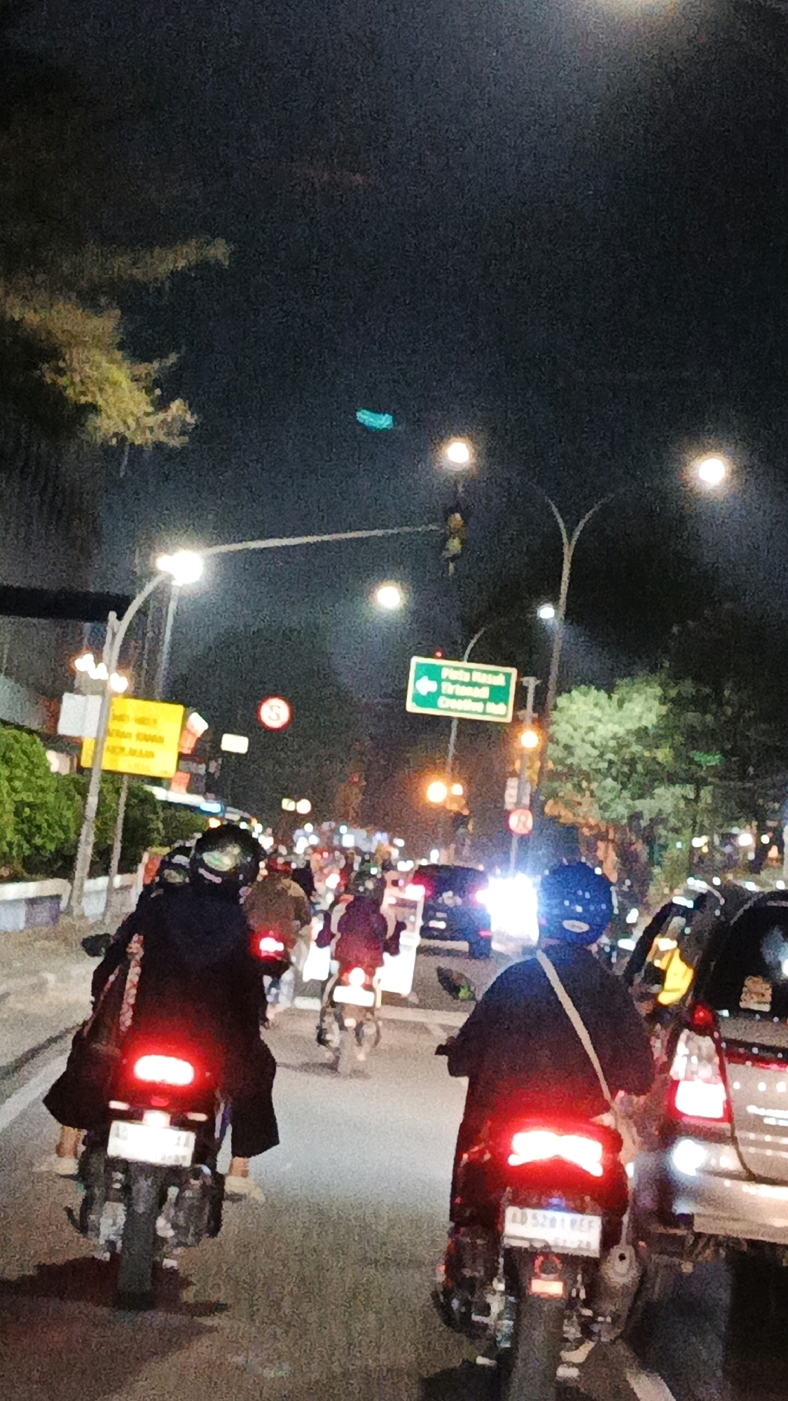 The image depicts a night scene of heavy traffic on a city street in Surakarta, Indonesia. Multiple motorcycles and cars are visible, with the taillights of the vehicles illuminating the road. A green sign with white text indicates "Pintu Masuk Tirtonadi Creative Hub." The sky is dark, suggesting it is nighttime, and streetlights are present. Several individuals are riding motorcycles, some wearing helmets. The overall atmosphere is one of a busy urban environment at night.