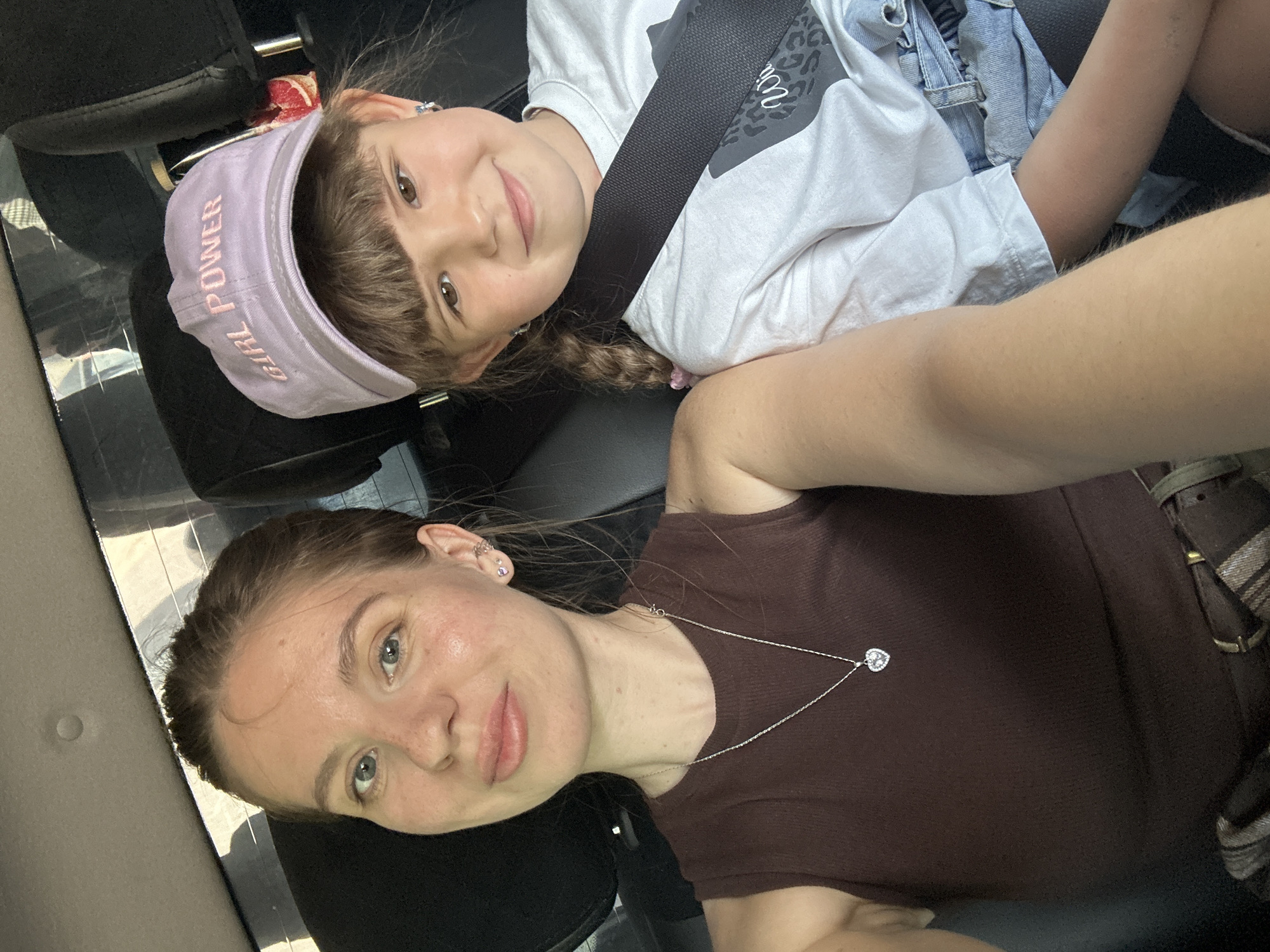 Two females are captured in a selfie within a vehicle. The younger female, positioned at the top of the frame, wears a lavender visor with "GIRL POWER" inscribed on it, along with a white shirt, denim overalls, and a seatbelt. Her hair is styled in a braid with a pink tie. The older female, at the bottom of the frame, has fair skin and wears a brown shirt and a silver heart pendant necklace. The vehicle's interior includes black seats and trim. The city of Unknown, Russia, cannot be confirmed from the image.