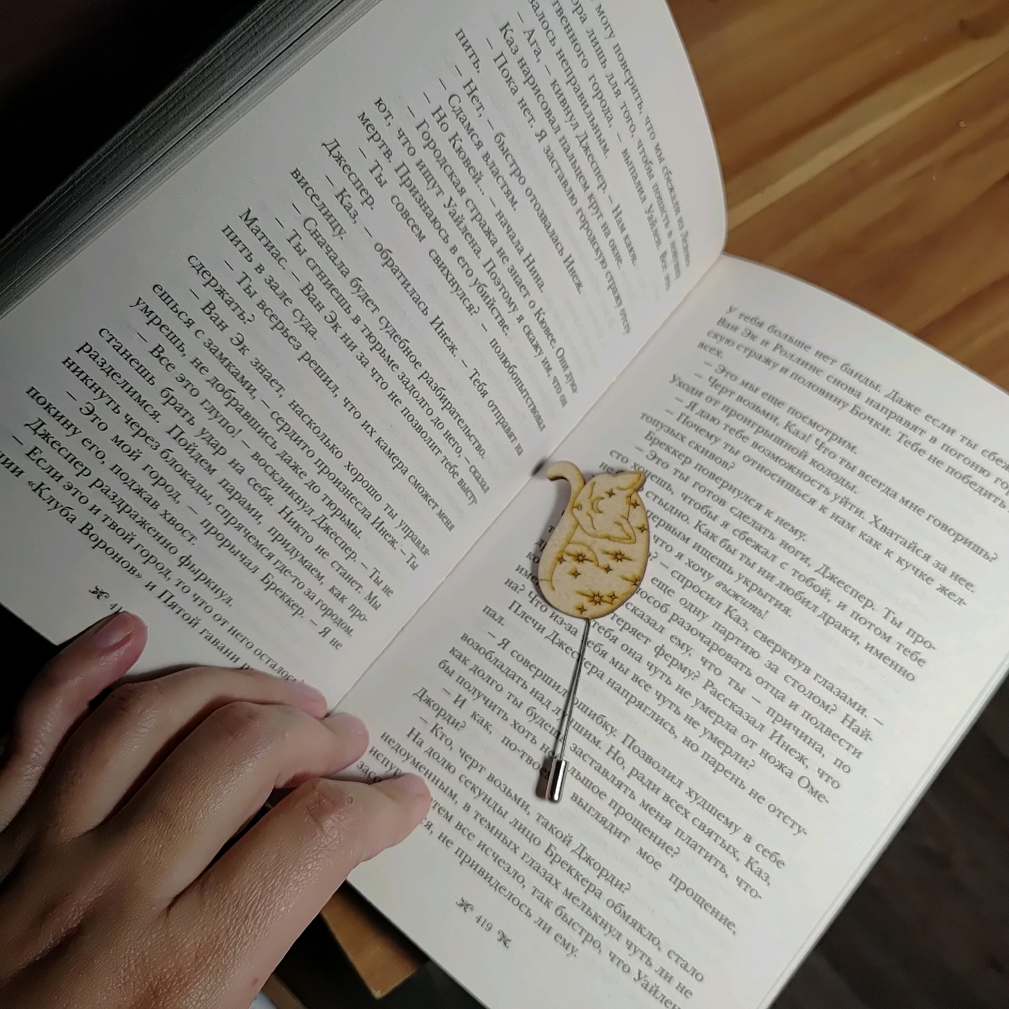 The image shows an open book with text in Russian. A wooden bookmark shaped like a cat with stars is placed on the page. A person's hand is visible on the left side of the image, holding the book open. The background is dark and out of focus. There is no information about the city of Марковский, Russia in the image.