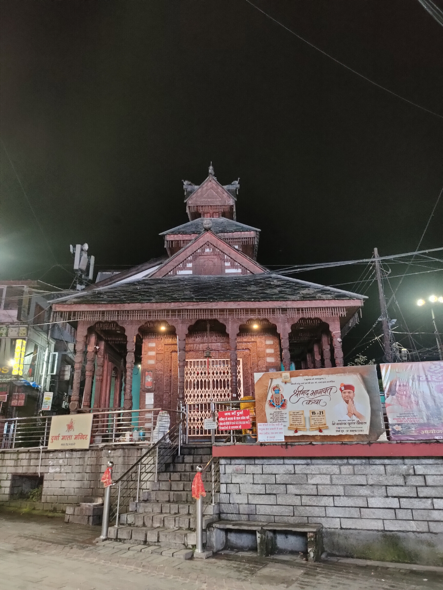 The image shows the Durga Mata Temple in Manali, India, at night. The temple is made of dark wood and has ornate carvings. It is lit by spotlights and streetlights. In front of the temple, there are posters advertising religious events. The temple is surrounded by brick walls and a set of stairs. A bench is visible in front of the temple. The sky is dark and clear, with a few wires and poles visible. The scene appears peaceful and festive.