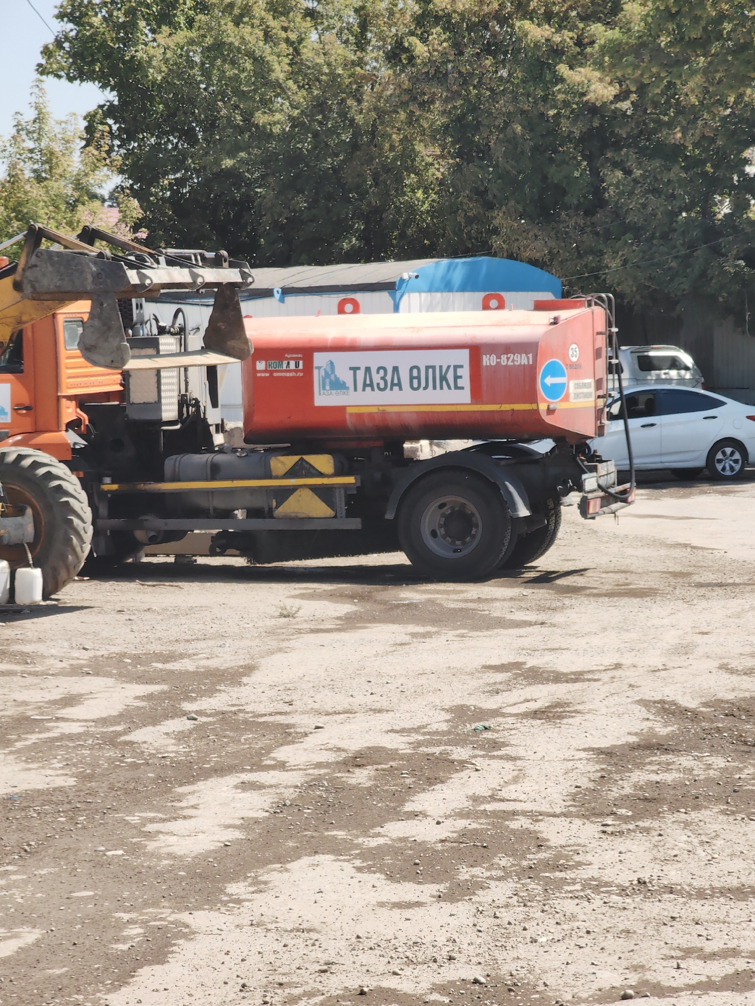 An orange excavator is parked next to a red tanker truck with "TАЗА ƏЛКЕ" written on its side in white letters. The tanker truck has a license plate that reads "KO-829A1". In the background, there is a white car and a building with a blue roof. The scene appears to be in an outdoor lot, possibly a construction site or a storage area, with a dusty ground. The overall environment is typical of an industrial or work area in a city.