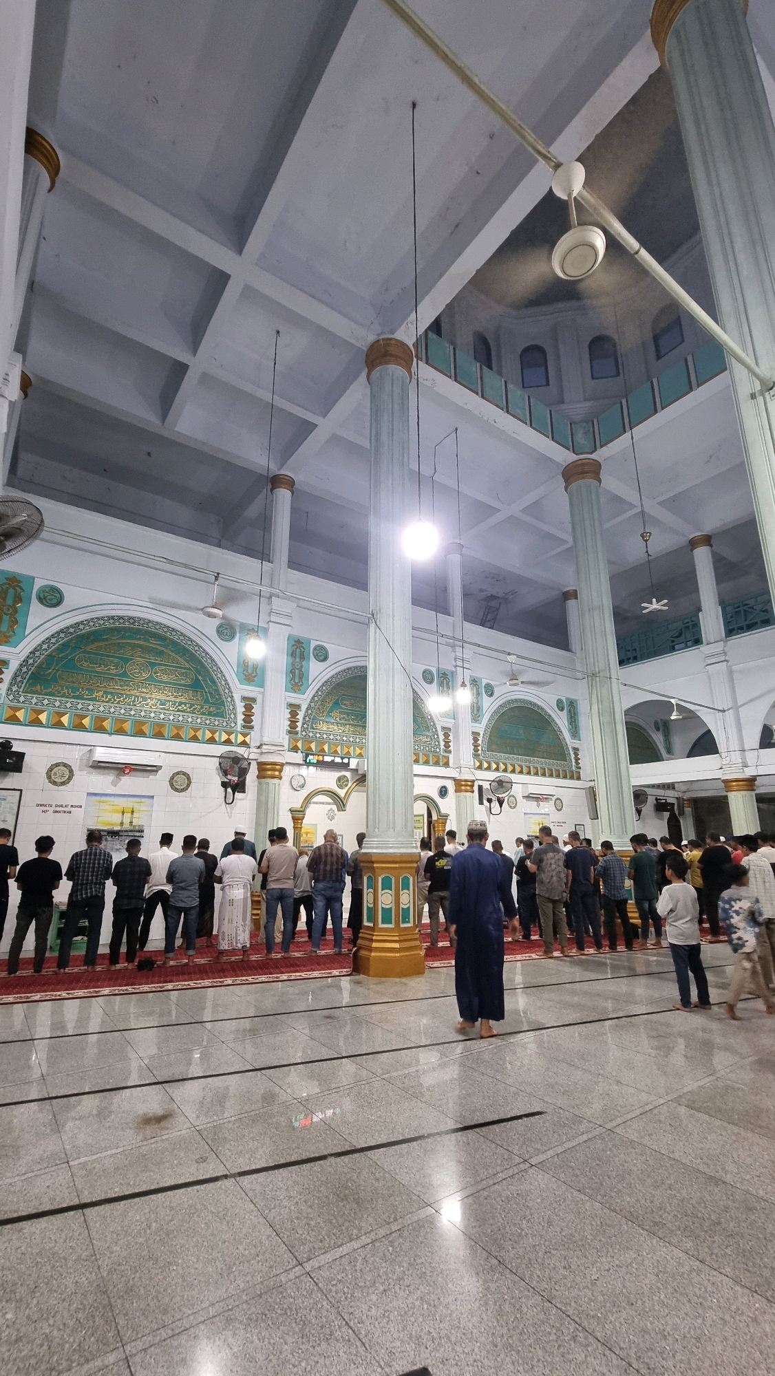 The image shows the interior of a mosque, with men standing in prayer. The mosque has high ceilings with a grid-like pattern and large, ornate columns. There are decorative arches along the walls with Arabic calligraphy. The floor is tiled and reflective. Several ceiling fans are visible. The scene appears to be in the city of Puloara, Indonesia, as indicated by signage visible in the image. The lighting is artificial, creating a bright interior.
