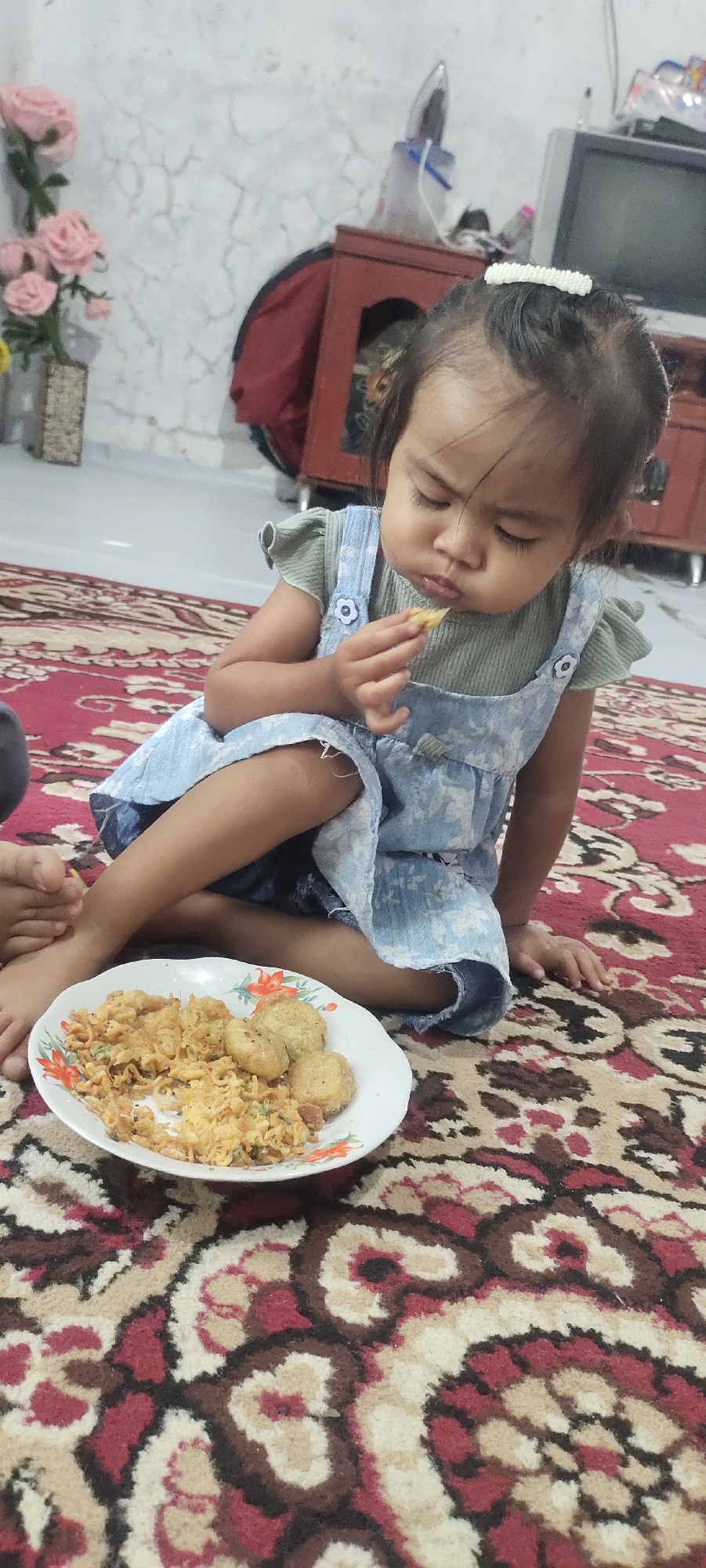 A young girl is sitting on a red patterned carpet in a room, eating from a plate of food. She is wearing a green top and a blue overall dress. Her eyes are closed as she brings a piece of food to her mouth. On the plate, there is rice and what appear to be fried balls. The background shows a wall with cracks, a piece of furniture, and some pink flowers in a vase. The scene appears to be in a domestic setting. The image does not provide specific information about the city of Cintadamai, Indonesia, or its urban features.