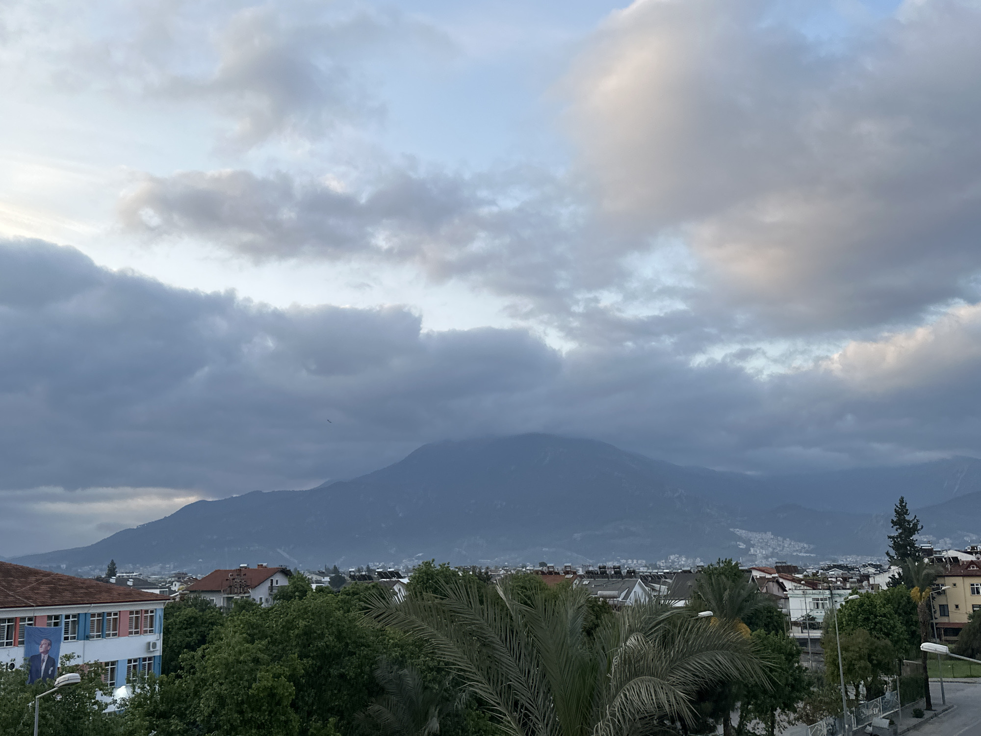 The image shows a cityscape in Fethiye, Turkey, under a cloudy sky. A large mountain dominates the background, its peak partially obscured by clouds. In the foreground, rooftops of buildings intermingle with green trees. A palm tree is visible in the lower center of the image. On the left, a building with a Turkish flag and an image of Mustafa Kemal Atatürk is visible. A street lamp is visible on the right side of the image. A small bird is visible in the sky.