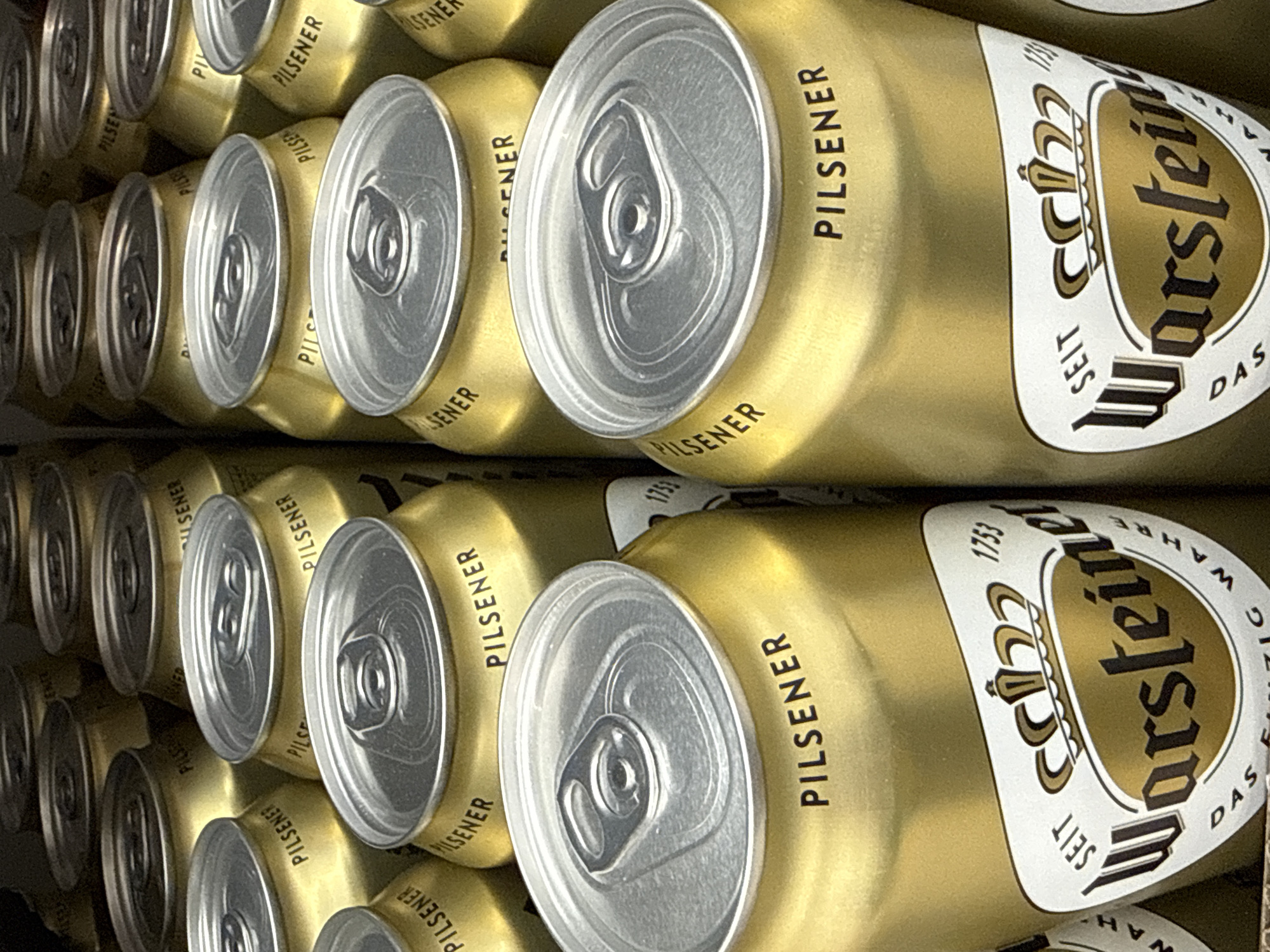 The image shows a close-up view of several golden-colored beer cans, specifically Warsteiner Premium Pilsener. The cans are arranged in rows, with the tops of the cans prominently featured in the foreground. The Warsteiner logo, which includes a crown and the text "Warsteiner", is visible on some of the cans. The cans are likely displayed for sale in a store or supermarket. The background is dark and out of focus, suggesting an indoor setting. There are no people or animals visible in the image. The scene is devoid of any specific location markers that would allow for pinpointing its presence in Bad Lauterberg im Harz, Germany.