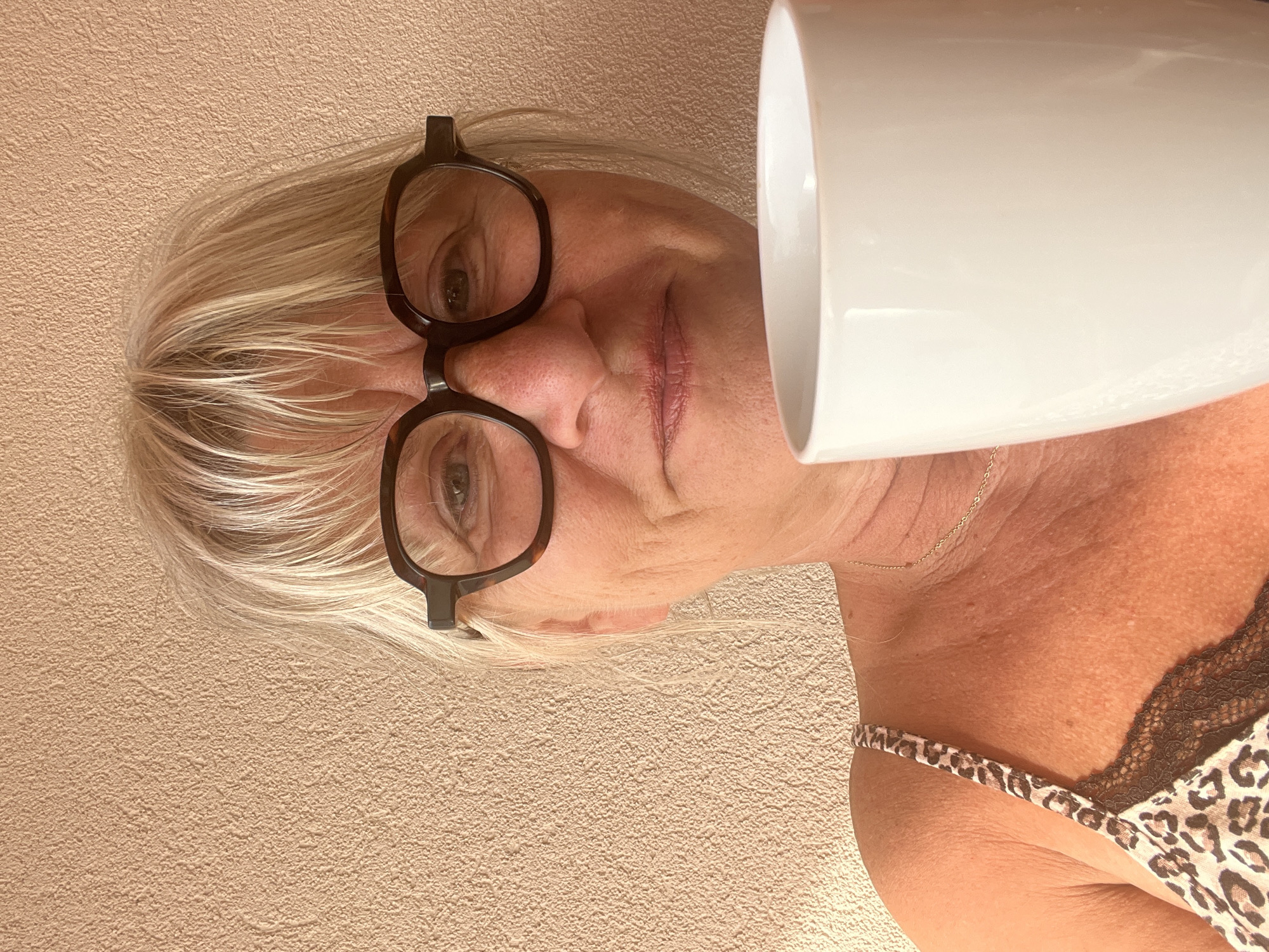 A woman with blond hair and glasses holds a white mug in front of her face. She is wearing a leopard print top with lace trim. The background is a textured, light brown wall. The scene appears to be indoors, possibly in a residential setting in Palavas-les-Flots, France, given the natural light and the casual attire.