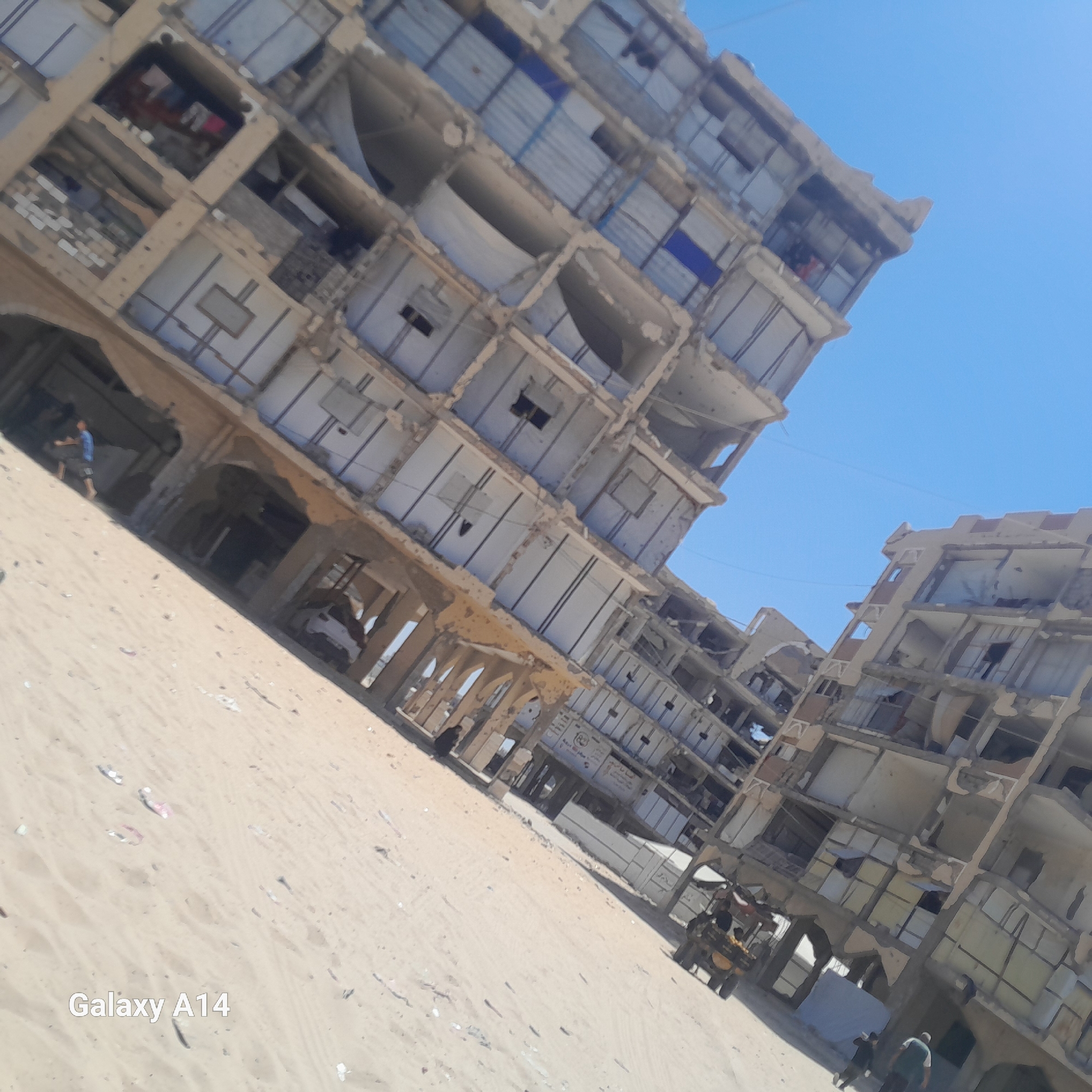 The image, taken in Khan Yunis, Palestinian Territories, shows a scene of significant destruction. Two partially collapsed multi-story buildings dominate the frame, their facades severely damaged, with exposed rebar, missing walls, and broken windows. The structures appear to be residential or commercial buildings, with visible signs of interior living spaces, including what looks like laundry hanging in one of the units. The ground is covered in sand and debris, suggesting a sandy environment or a location near a beach. In the foreground and midground, several individuals are visible. One person is standing near the entrance of one of the damaged buildings, while another is seen further in the distance. A small cart or vehicle is also present, with at least one person on it. The sky is clear and blue, providing a stark contrast to the devastation of the buildings. The overall impression is one of a recently impacted area, with the architecture and the presence of people indicating ongoing life amidst the ruins.