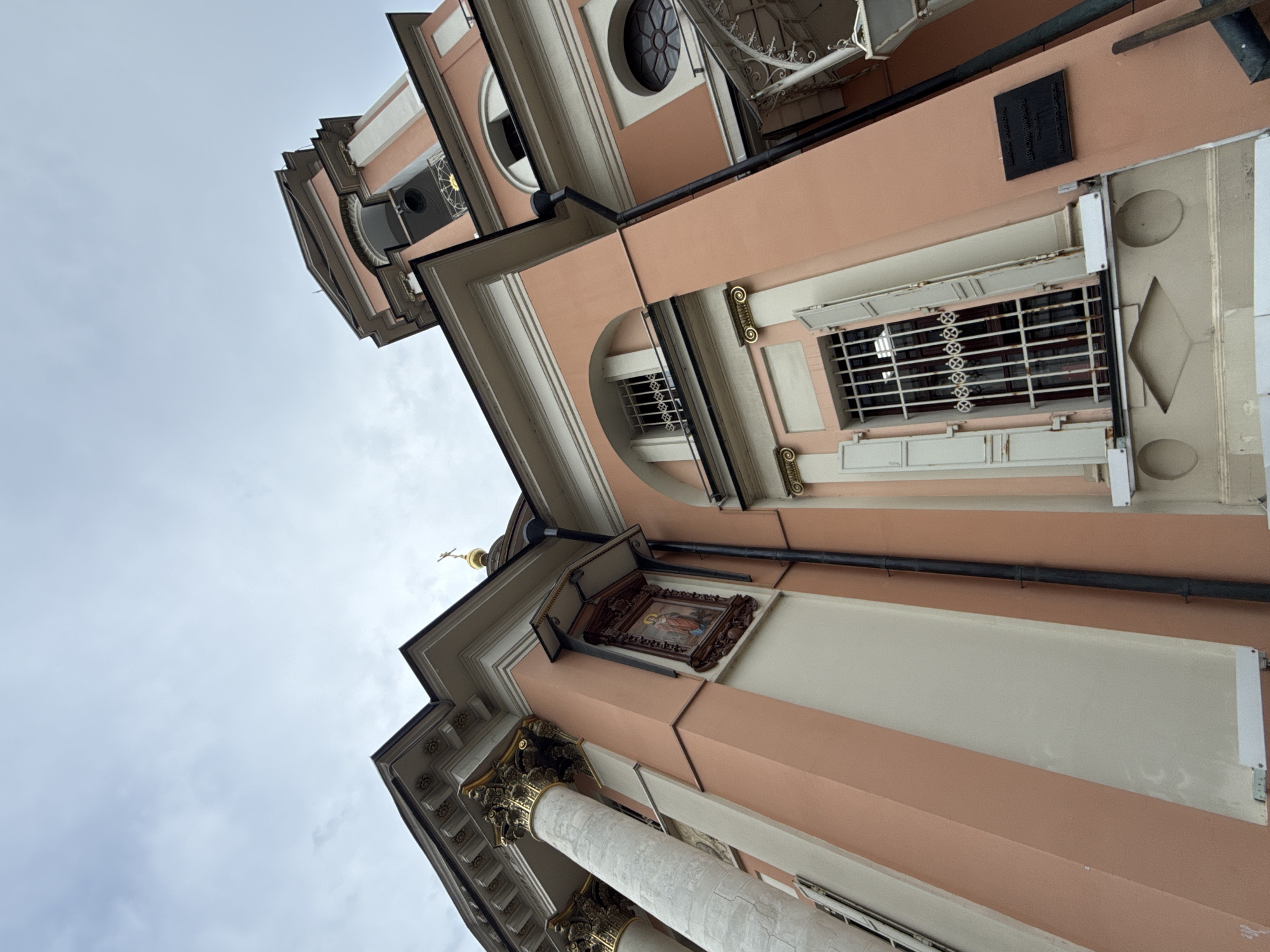 This is a low-angle, ground-level shot of the Church of Saint Barbara, located on Varvarka Street in Moscow, Russia. The church is painted a pale pink with cream-colored accents and features classical architectural elements such as columns and ornate cornices. A bell tower rises in the background. The sky is overcast. There are no people visible in the image.