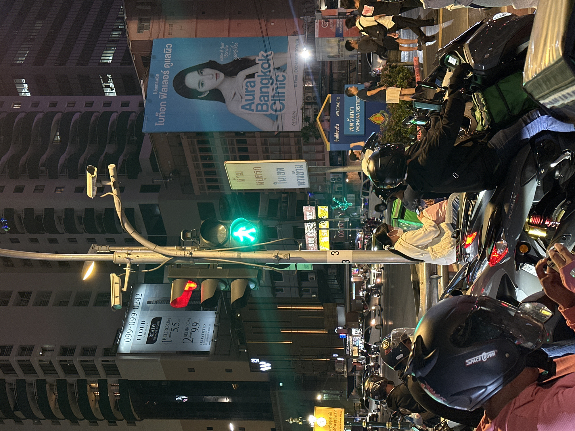 The image shows a street scene at night in the Vadhana district of Bangkok, Thailand. The traffic signal displays a green pedestrian crossing symbol. A line of motorcycles is waiting at the intersection. Close to the foreground, a person is holding a phone, possibly taking a picture. In the background, a large billboard advertises "Aura Bangkok Clinic" with the image of a woman. Another advertisement displays contact information for apartments at Cloud Sukhumvit 23. Street signs indicate "Vadhana District" and warnings related to construction. People are also walking on the sidewalk.