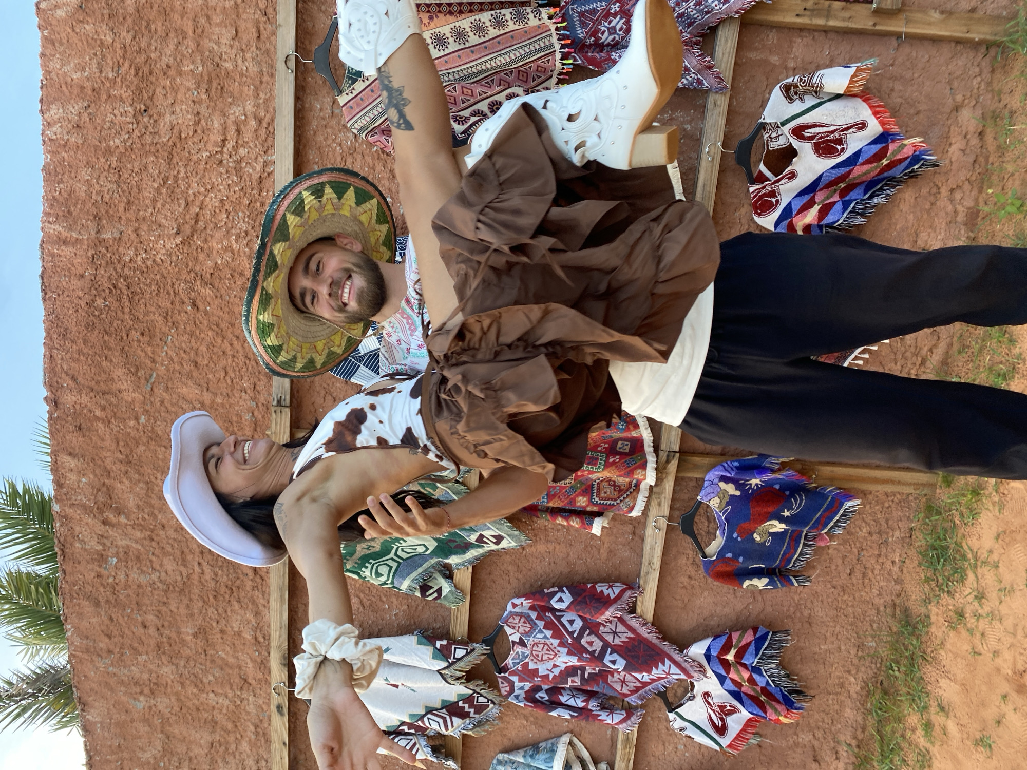 A man in a sombrero and patterned shirt is holding a woman in a cowboy hat and cow-print top. The woman is wearing a brown ruffled skirt and white cowboy boots, and her legs are extended. They are posing in front of a wall of colorful woven ponchos hanging on wooden racks. The wall appears to be made of reddish-brown stucco. Palm fronds are visible at the top of the image, suggesting an outdoor location with a warm climate. The ground in the foreground is sandy and has some sparse green vegetation. The scene has a festive, possibly tourist-oriented atmosphere, and the attire suggests a theme of Western or Mexican culture. Given the context, the location is likely a market or a stall selling souvenirs in Mui Ne, Vietnam.