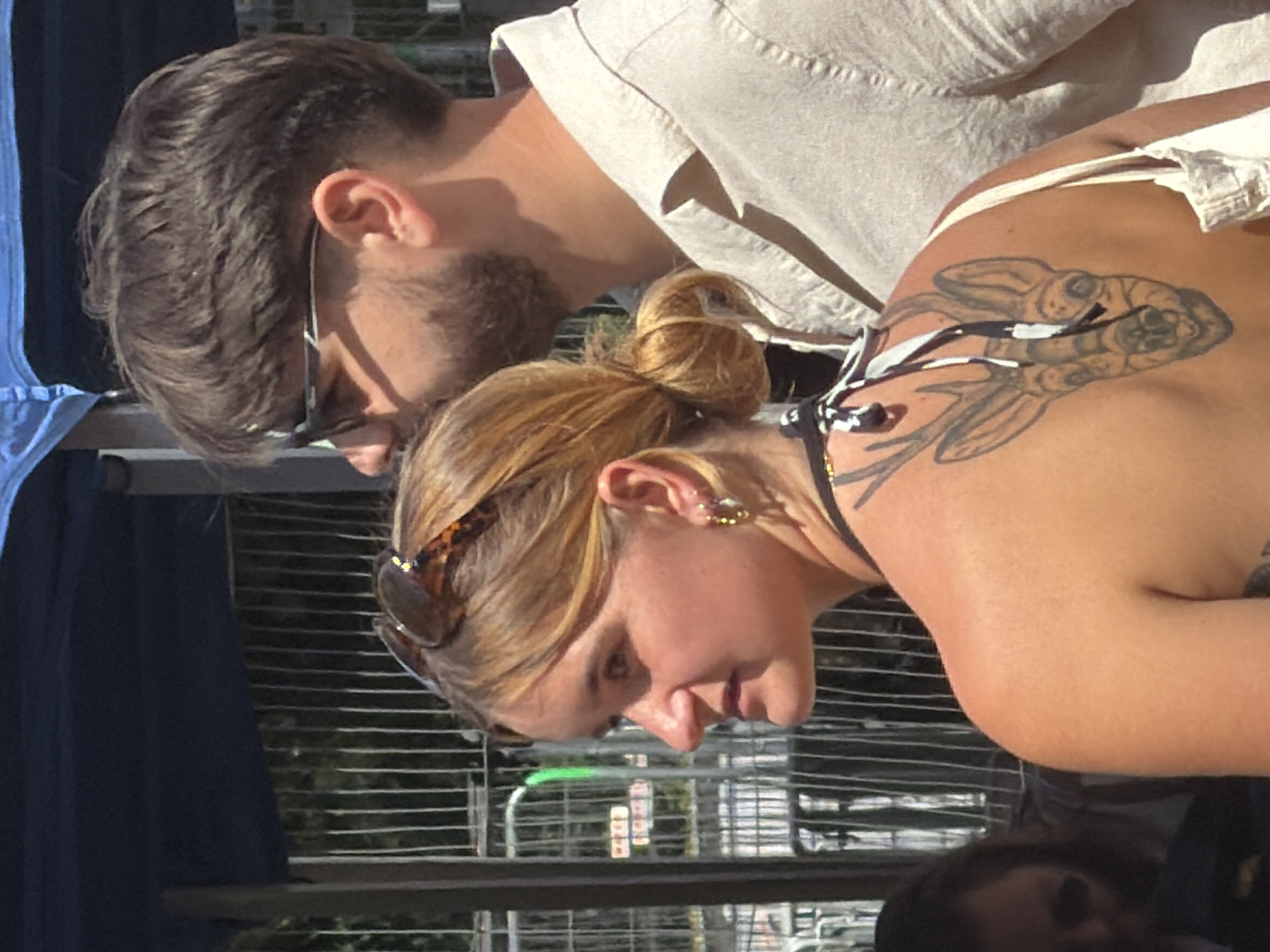 This is a medium shot, captured outdoors on a sunny day, focusing on two individuals in the foreground and a third partially visible in the background. The setting appears to be an outdoor event or gathering, with a blue canopy and metal fencing visible behind the subjects.
In the foreground, a woman is positioned to the left, her body turned to her left and her head looking left. She has blonde hair tied back in a bun, and she is wearing large hoop earrings. A tattoo of a deer's head is visible on her upper back. A man is to her right, facing primarily forward with his head turned slightly to his left, looking in the same direction as the woman. He has short brown hair and a beard, and he is wearing sunglasses.
The woman is wearing a black and white patterned bikini top. The man is wearing a light-colored, button-down shirt. The man in the background is partially obscured and appears to be wearing sunglasses. The overall lighting suggests bright daylight. The location is stated to be Esch-sur-Alzette, Luxembourg, and the background elements such as the fencing and the general atmosphere suggest a public gathering or festival.