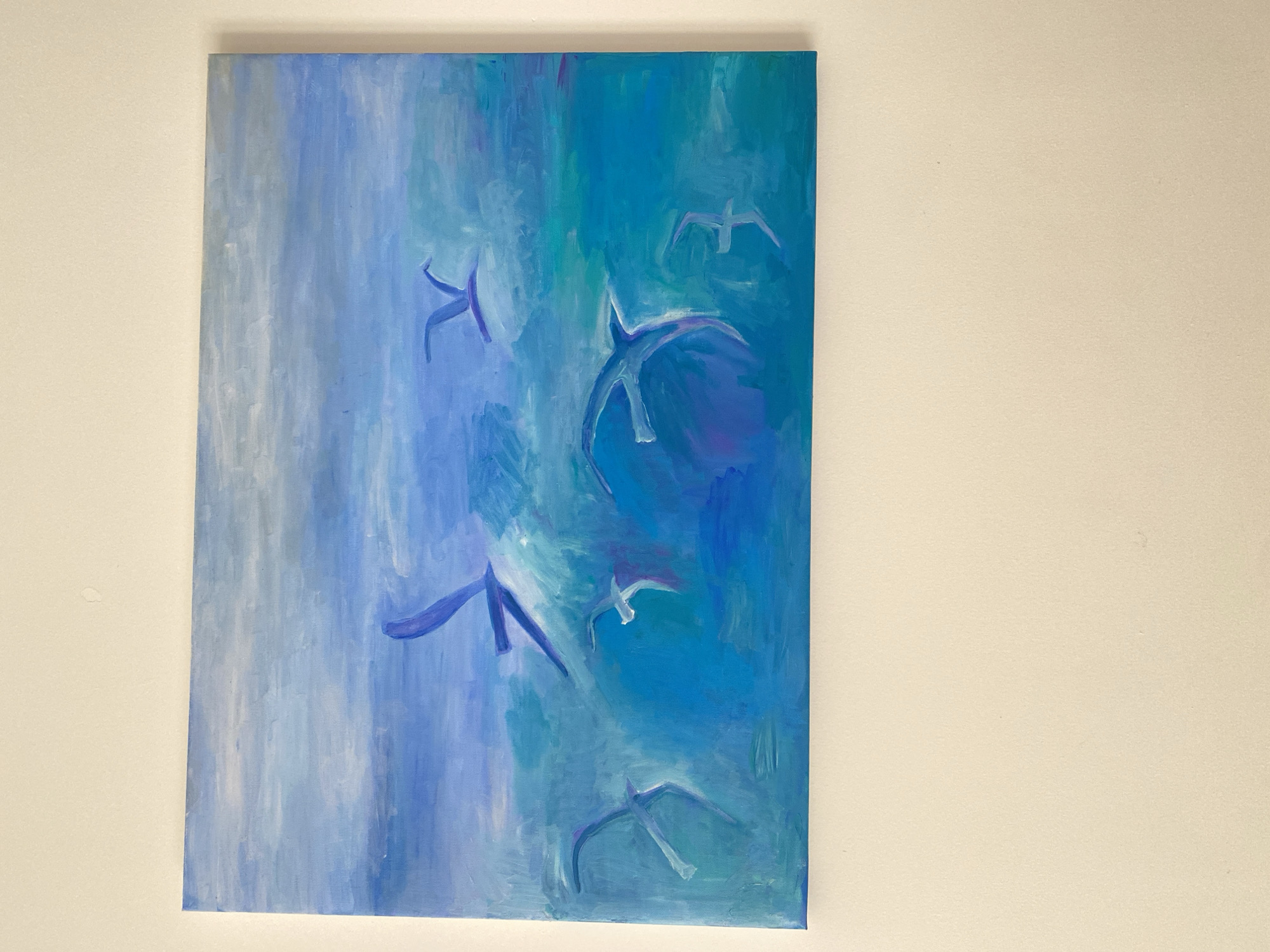 A painting of birds in flight hangs on a light beige wall. The painting is a vertical rectangle and features an abstract depiction of birds with blue and white brushstrokes. The background consists of vertical gradations of blues and whites. There is no evidence this image was taken in Lâm Viên - Đà Lạt, Vietnam.