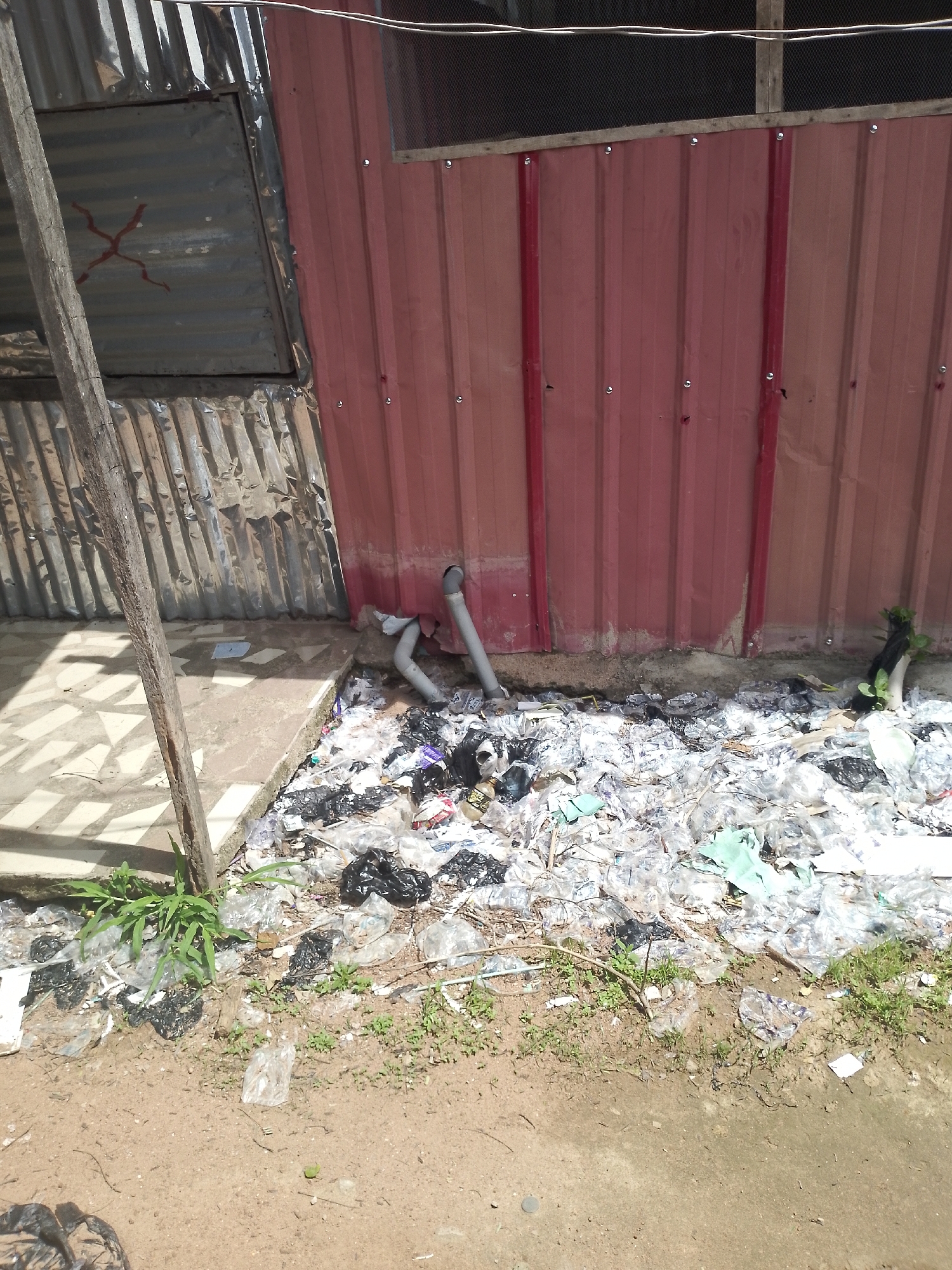 The image depicts an outdoor scene in Jalingo, Nigeria, characterized by significant litter and a weathered corrugated metal structure. In the foreground, the ground is a mix of dirt and scattered debris, including plastic bags and wrappers, with a few small green plants growing amidst the waste. A discarded plastic bag is visible in the lower left corner. The midground reveals a large pile of trash, primarily composed of translucent and black plastic bags, some containing visible contents, along with other assorted refuse. Two gray PVC pipes extend from the wall into the trash. Behind the trash is a wall constructed of red corrugated metal panels, showing signs of wear and dirt. To the left of the red wall, a section of silver corrugated metal structure is visible, with a wooden support post in front of it. This silver section appears to have some kind of panel or door, marked with a red 'X' shape. The overall environment suggests an unkempt urban or peri-urban area.