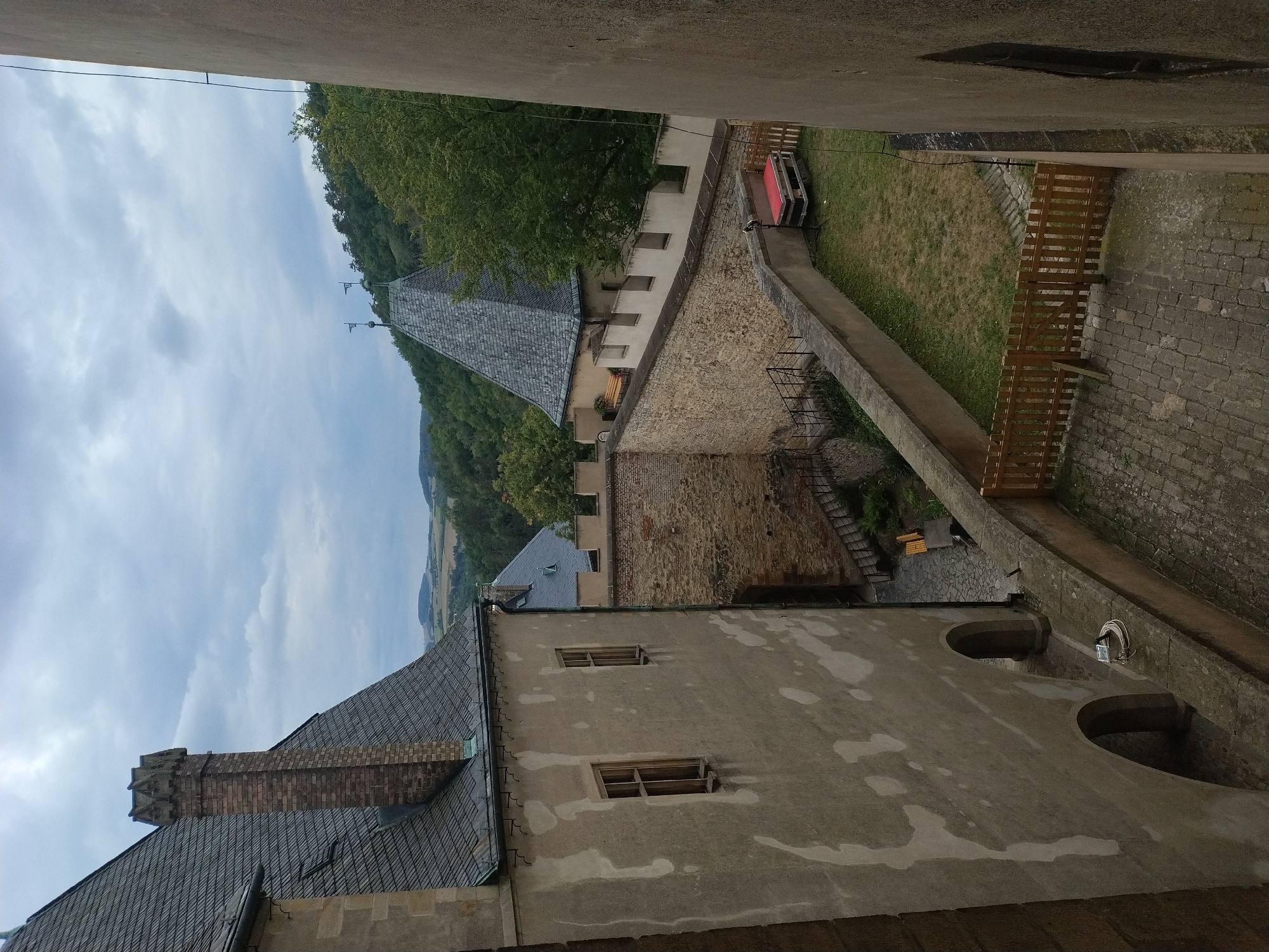 The image depicts a view from a higher vantage point within the Karlštejn Castle in Karlštejn, Czechia. In the foreground on the left is the weathered, pale facade of a castle building with two rectangular windows, one slightly ajar. To the right of this building, a stone wall with crenellations runs horizontally across the frame. Beyond this wall, a grassy courtyard is visible, with a wooden fence bordering a cobbled area. A bright red, pallet-mounted structure sits in the courtyard. In the background, a steep, wooded hillside rises, topped with trees and a conical-roofed structure with two flags. The sky above is overcast with heavy clouds. The scene appears to be an outdoor area within the castle grounds, likely a courtyard or a section of the defensive walls.