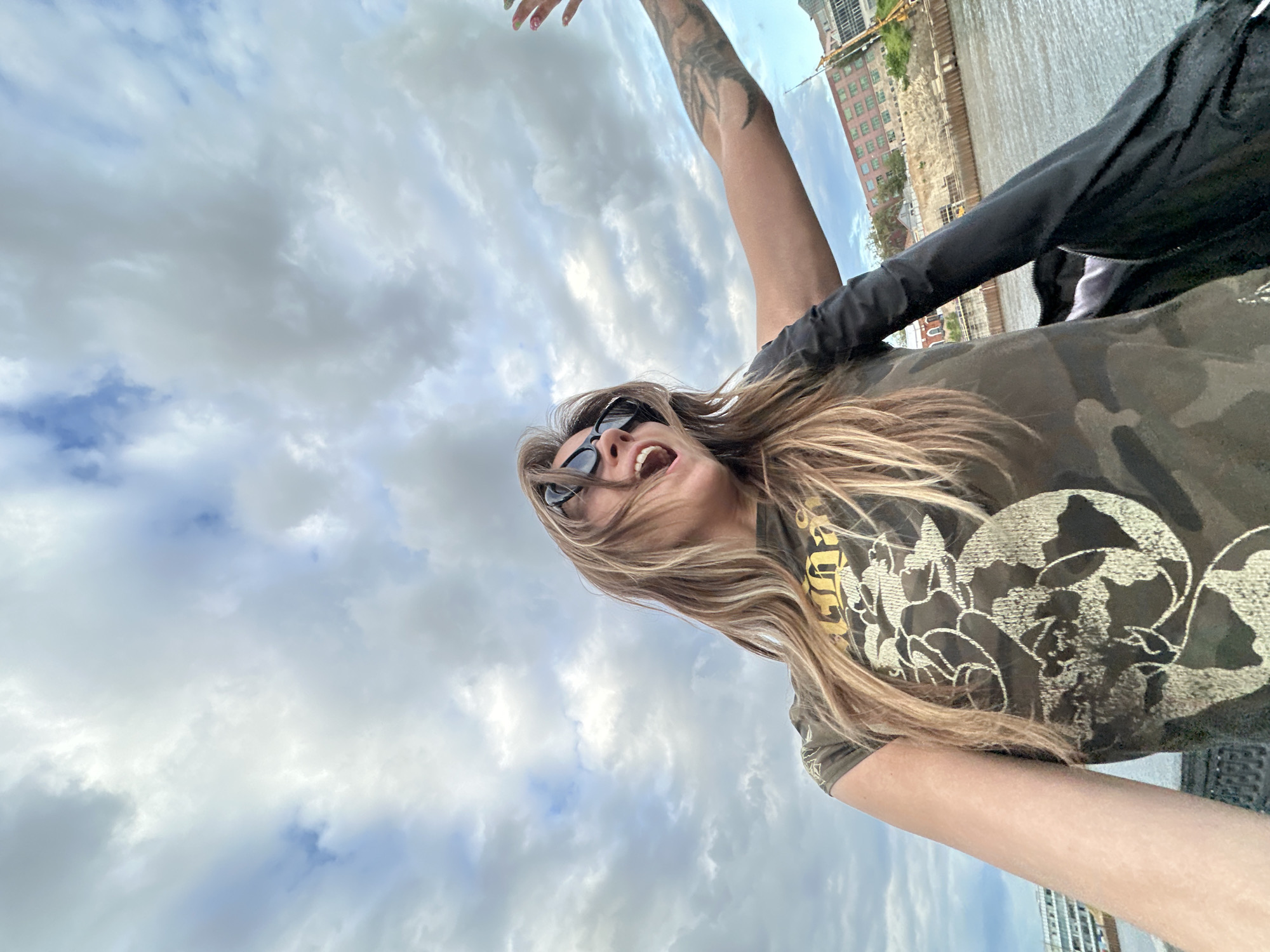 A low-angle, full-body shot captures a young woman from the chest up, posing for a selfie outdoors. She stands on a body of water, with a cityscape and cloudy sky in the background. The woman is wearing a camouflage t-shirt, black sunglasses, and has long, blonde hair. Her mouth is open as if speaking or singing, and her right arm is raised and extended, with her hand out of frame. A black jacket is draped over her right arm, partially obscuring her body. The cityscape features a body of water, a waterfront with buildings under construction, and a hazy sky filled with scattered clouds.