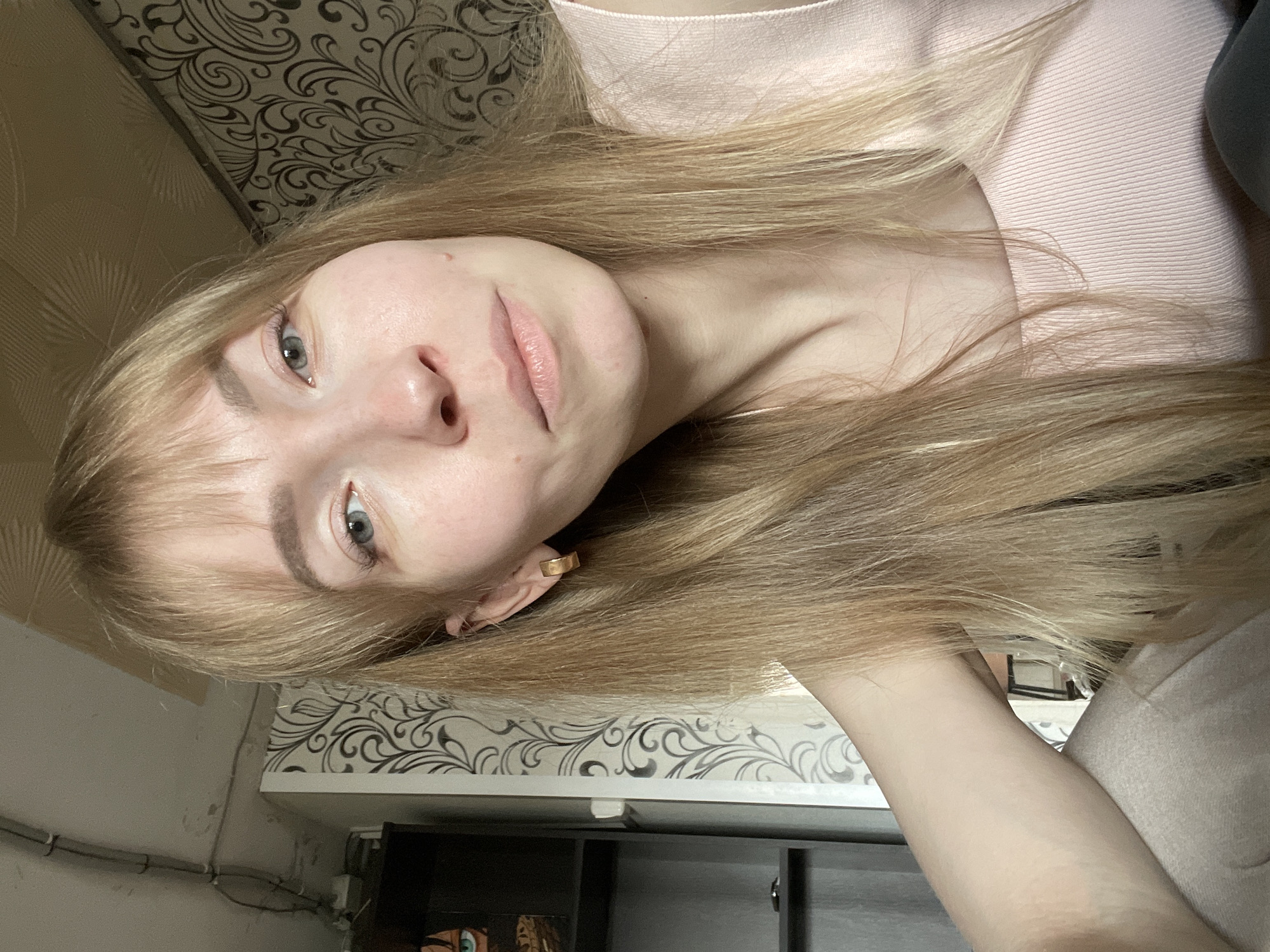 A young woman with blonde hair and fair skin is positioned in the center of the frame. She is wearing a light pink top. Her facial features are prominent, including light-colored eyes and full lips. She is wearing a gold earring in her left ear.
The background features a wall with patterned wallpaper. Beneath the wallpaper is a wall shelf with cords and wiring. A dark-colored cabinet is located under the shelf. The scene appears to be indoors, possibly in a residence. The image does not contain enough data to identify the location in Perm, Russia.