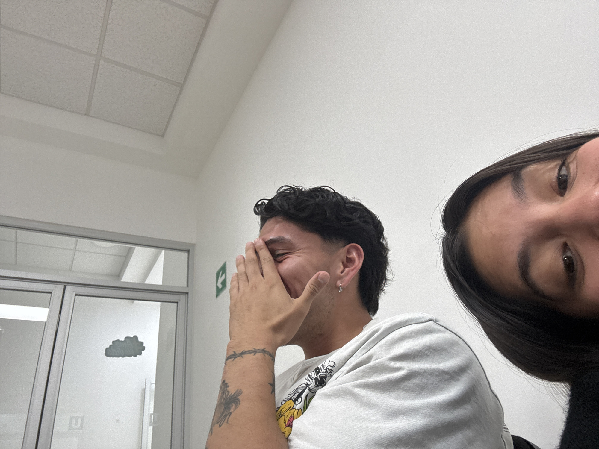 The photo shows two people in what appears to be an indoor space, likely an office or institutional setting, evidenced by the ceiling tiles, plain white walls, a door with a cloud-shaped decoration, and an exit sign. The man on the left is covering his face with his hand, smiling. He has short dark hair, an earring, and tattoos on his arm. He is wearing a white t-shirt with a graphic design. The second person, positioned on the right side of the photo, has dark hair and is looking down and slightly to the left. The overall impression suggests that the photo was taken in a university setting in Puebla, Mexico.