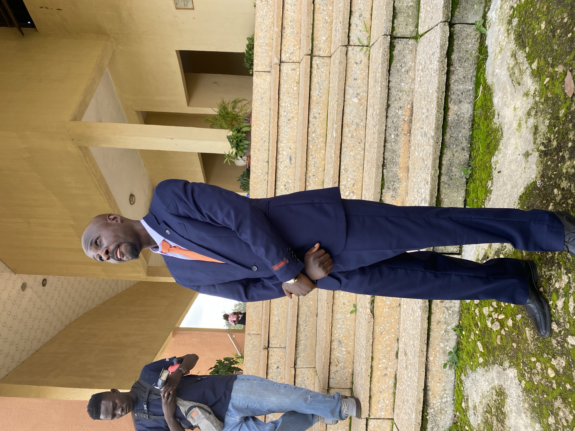 The media file depicts two individuals outdoors, possibly in Nigeria, given the location context. The main subject is a bald Black man wearing a navy blue suit, an orange tie, and black dress shoes. He is standing and looking to his left, with his hands clasped in front of him. He appears to be in his 40s or 50s and has a goatee. He has a serious expression. Behind him, a younger Black man is partially visible, wearing a navy blue t-shirt, blue jeans, and grey sneakers. He is holding a phone and a bag, and appears to be looking down.
The scene is set against a backdrop of a building with a yellow facade and a staircase made of large, textured concrete blocks. Moss and small plants are growing on the steps and the ground to the right, suggesting a humid or damp environment. The lighting suggests it is daytime, possibly overcast due to the diffused light. There is a small sign on the wall of the building with some text, but it is not legible in the image. The overall impression is that of a formal or semi-formal gathering, or perhaps a casual encounter.