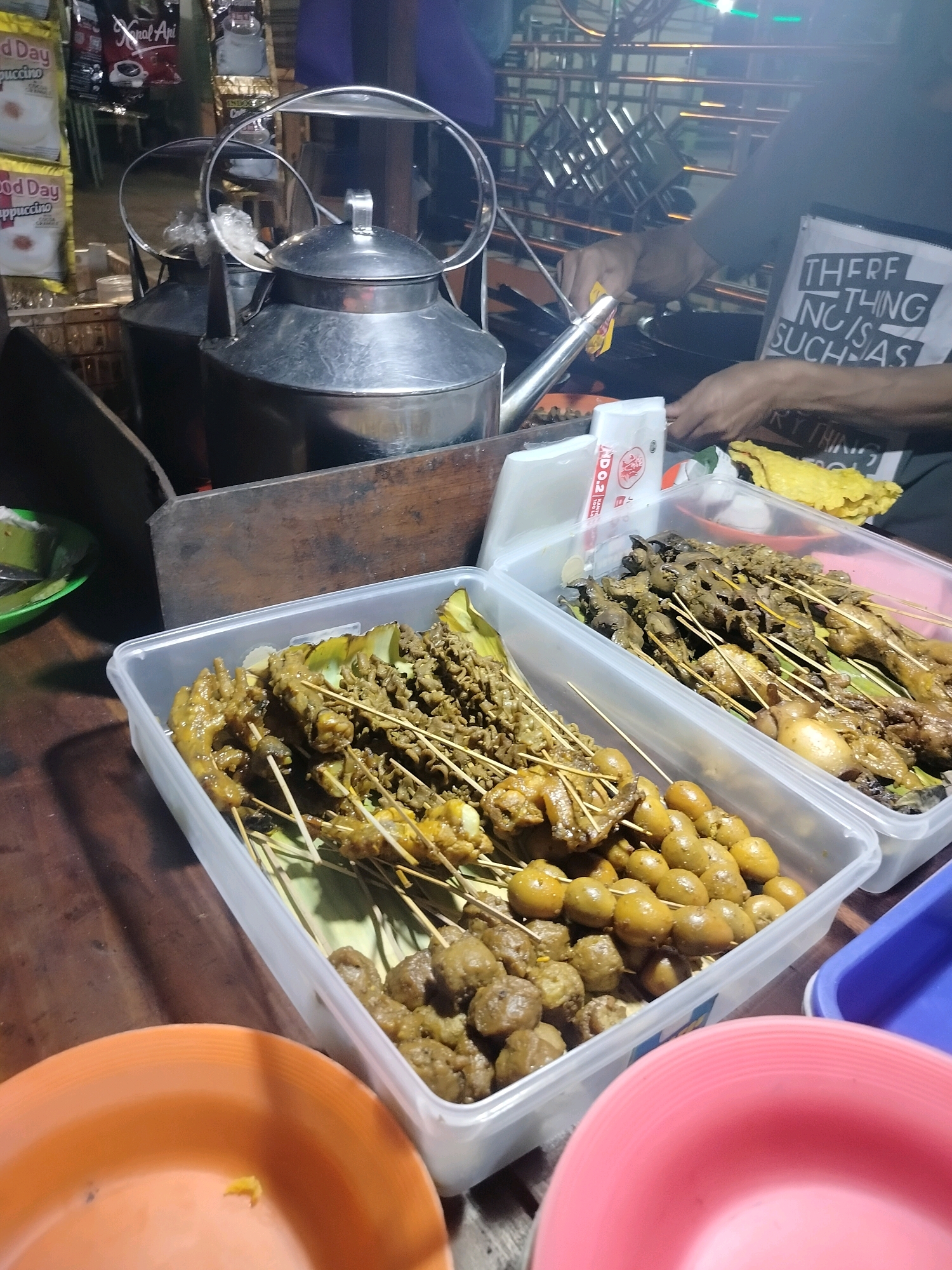 The image captures a street food stall in Kroya, Indonesia, at night. In the foreground, two transparent plastic containers are filled with various skewers of food, including what appear to be chicken feet, quail eggs, and various meat cuts. One container is in focus, showing the food items clearly. To the left of the containers, an orange plastic plate is visible. In the background, two large metal kettles are present, suggesting a beverage is being prepared or served. A person's hand is seen reaching towards the kettles and a cooking surface, indicating preparation is underway. A white t-shirt with text that reads "THERE IS NOTHING SUCH AS" is worn by the person. Packets of instant coffee and other items are displayed on a rack behind the kettles. The environment appears to be an outdoor setting, likely a street market or roadside stall, given the ambient lighting and the nature of the food being sold.