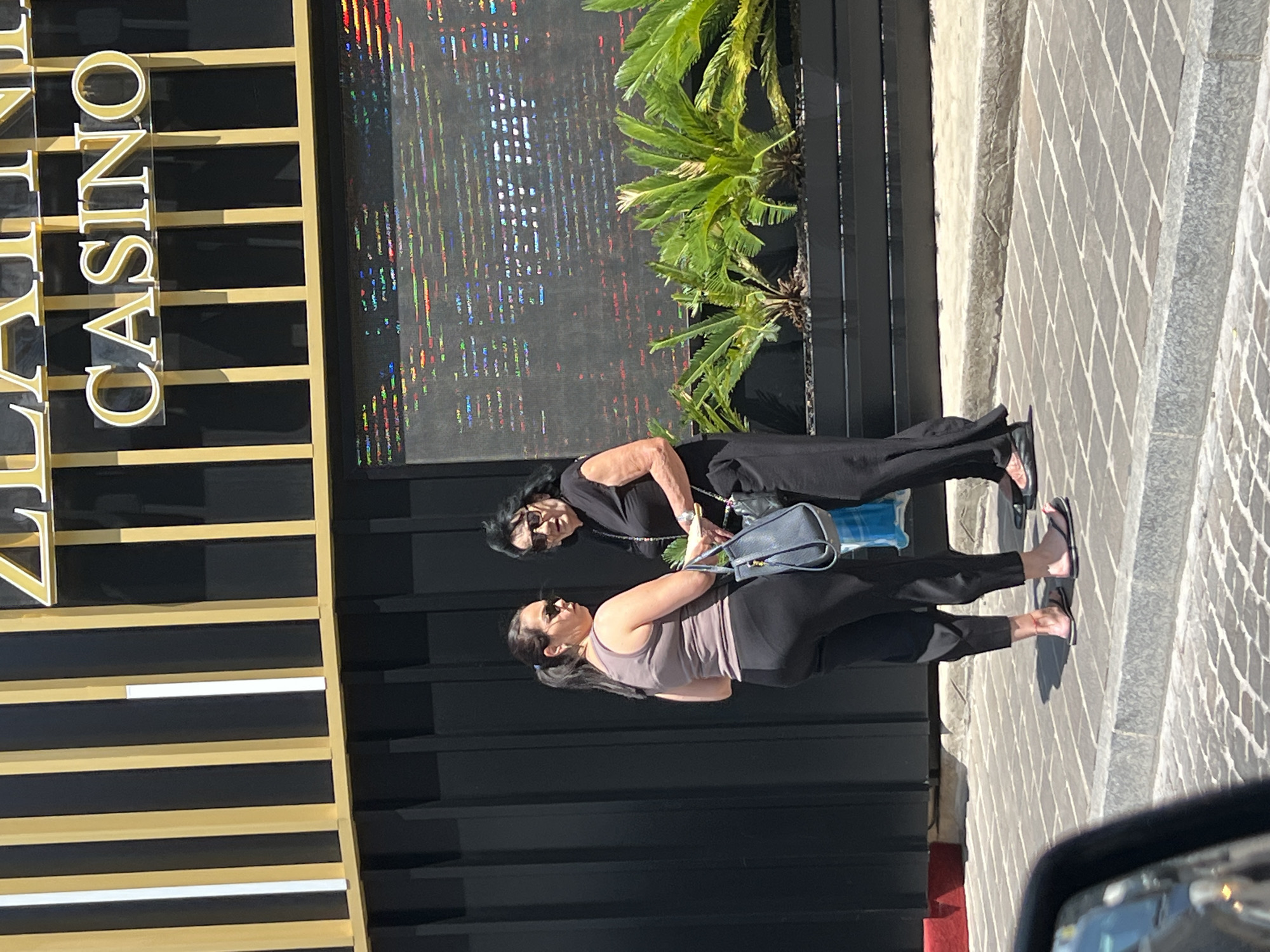 Two women are standing on a sidewalk in front of a building with a sign that reads "CASINO". The woman in the foreground is wearing a grey tank top and black pants, and she is looking to her left. The woman in the background is wearing a black top and black pants, and she is looking to her right. She is holding a blue bag and a green plant. The sun is shining, suggesting it is daytime. The building has a modern design with a facade featuring horizontal black and gold elements, and a digital screen displaying colorful abstract patterns. The sidewalk is paved with grey and white tiles laid in a diamond pattern. In the foreground, a car's side mirror is partially visible, indicating the photo was taken from inside a vehicle. The scene appears to be in a public area, possibly a tourist destination given the signage and architecture.