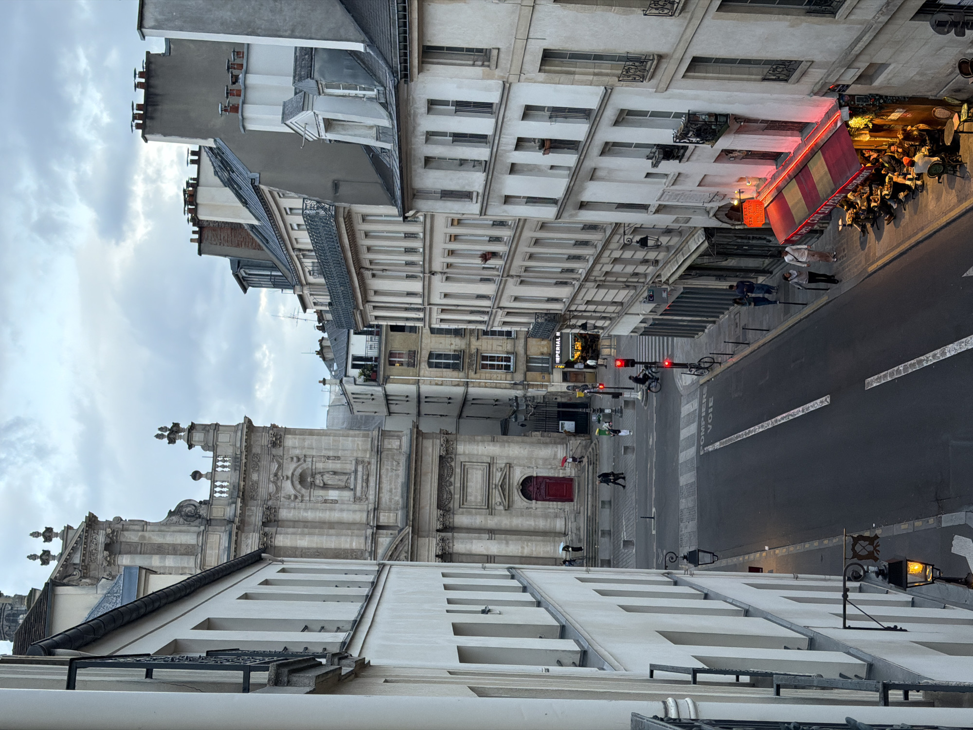 This image captures a street scene in Paris, France, viewed from an elevated position. The foreground features a section of a building, possibly the location of the photographer. Below, a street stretches into the distance, flanked by tall, ornate buildings characteristic of Parisian architecture.
People can be observed walking along the sidewalks and crossing the street. A cyclist is also visible on the road. A restaurant with an awning occupies a prominent position on one side of the street, with patrons seated at tables, suggesting an active street life. Traffic lights are present, indicating vehicular traffic.
The buildings display a variety of architectural details, including windows, balconies, and decorative elements. A church or another religious structure is also visible, featuring a facade with statues and ornate carvings. The sky is overcast, suggesting a cloudy day.